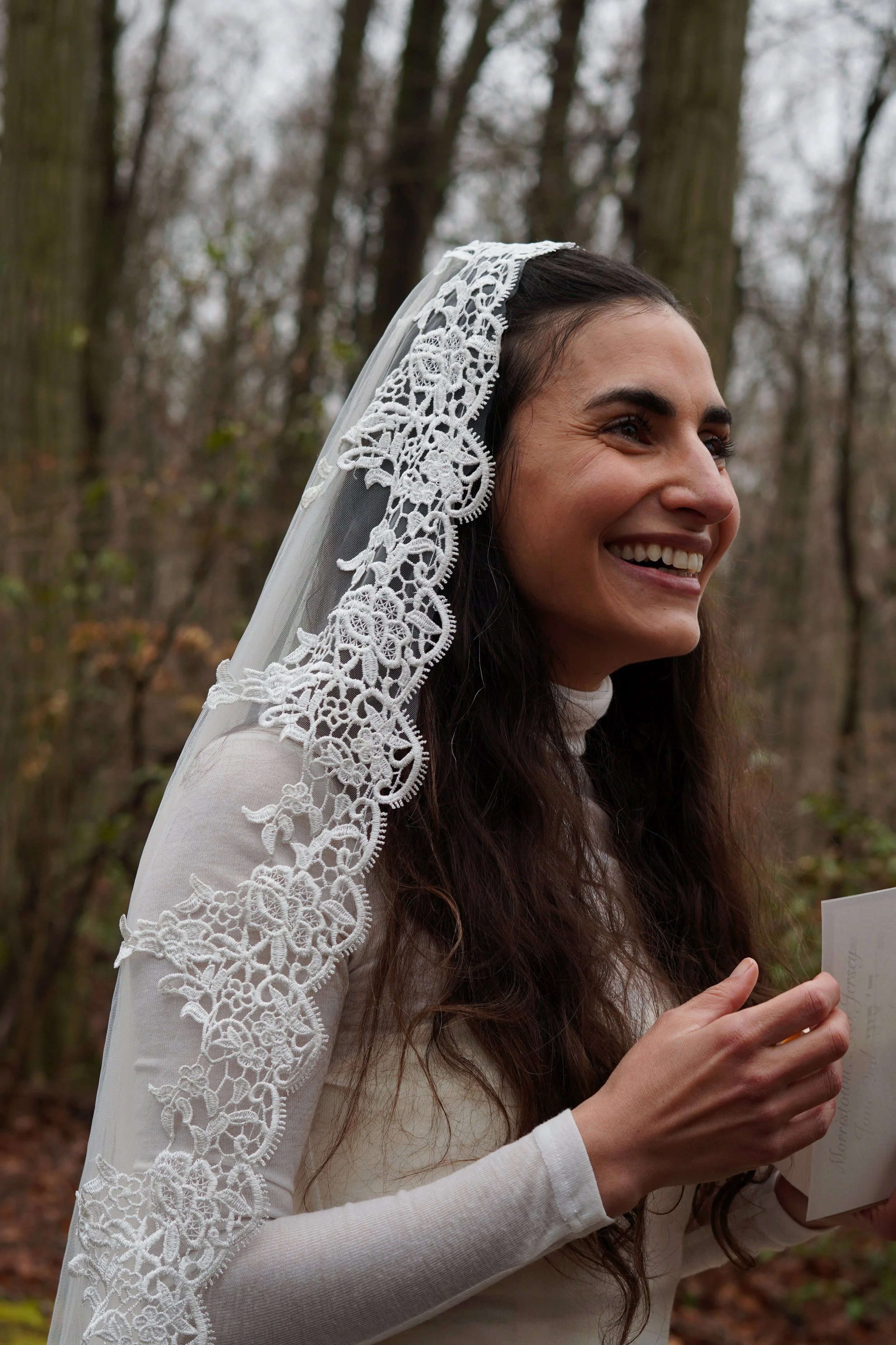 A woman smiling outdoors in a wooded area, wearing a wedding veil with lace on her head and holding a card. Cheap wedding photographer northern nj