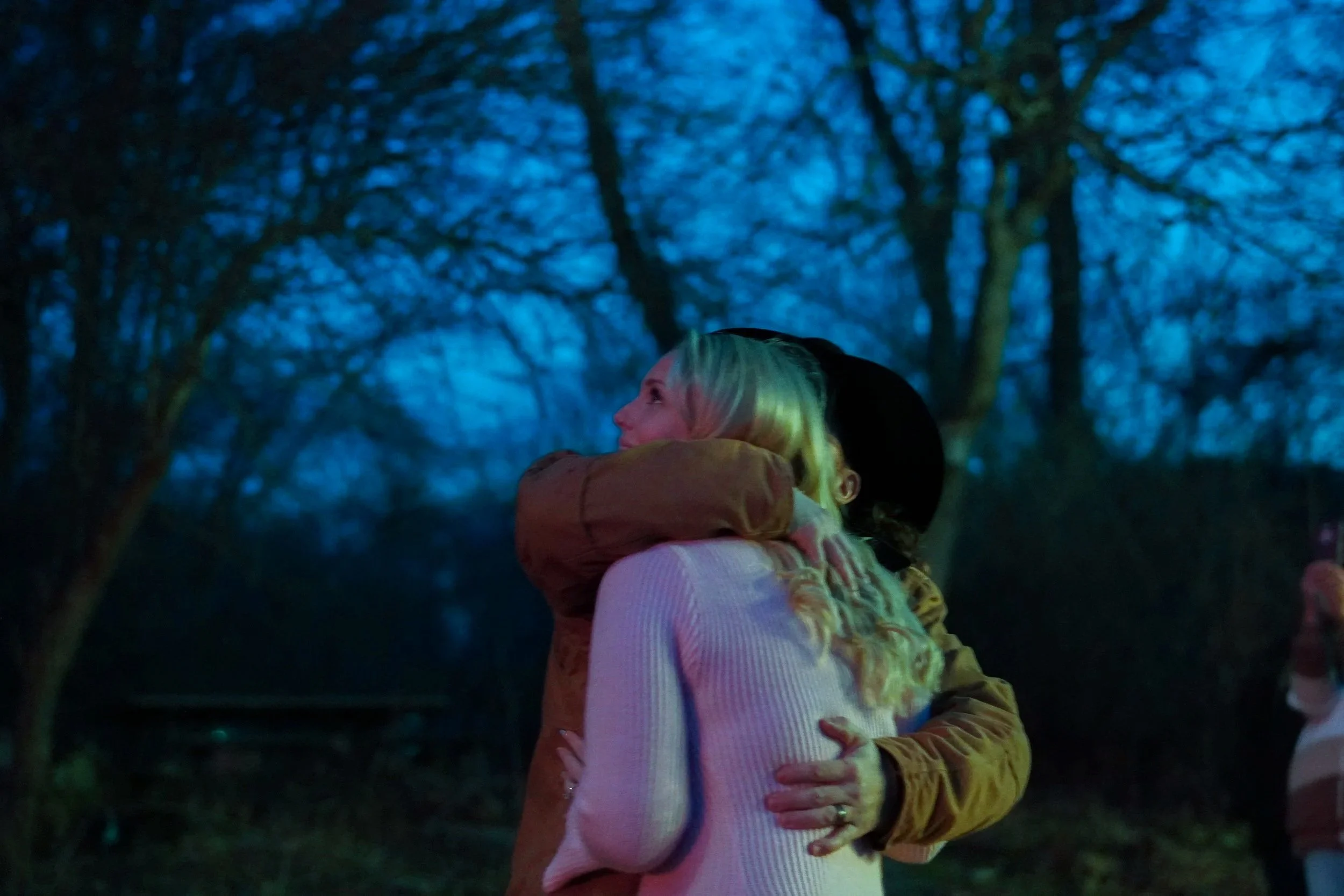 Two women hugging outdoors at dusk, trees in the background. Gender announcement photos.