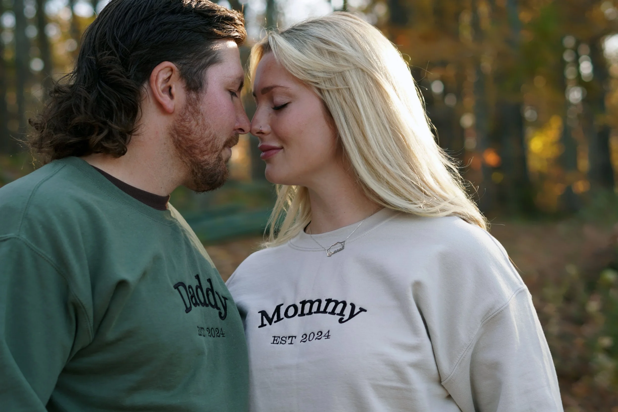 A couple standing close with foreheads touching in a forest during autumn, both wearing sweatshirts that say 'Daddy' and 'Mommy' with 'EST 2024' underneath. Baby announcement photos