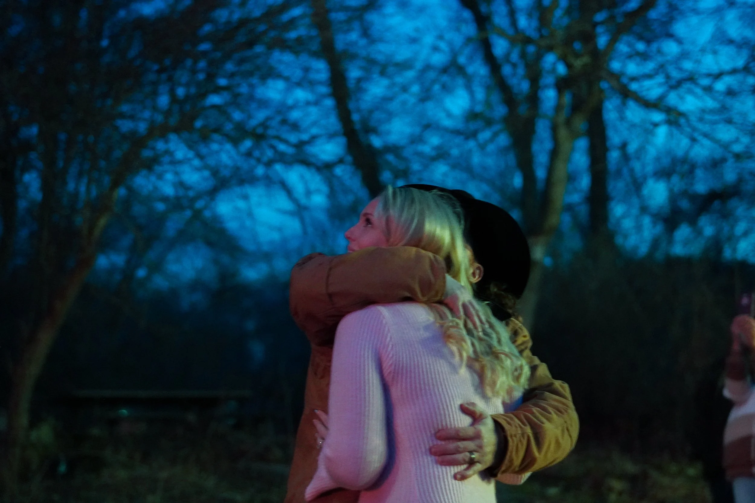 Two women hugging outdoors at dusk, trees in the background. Gender announcement photos.