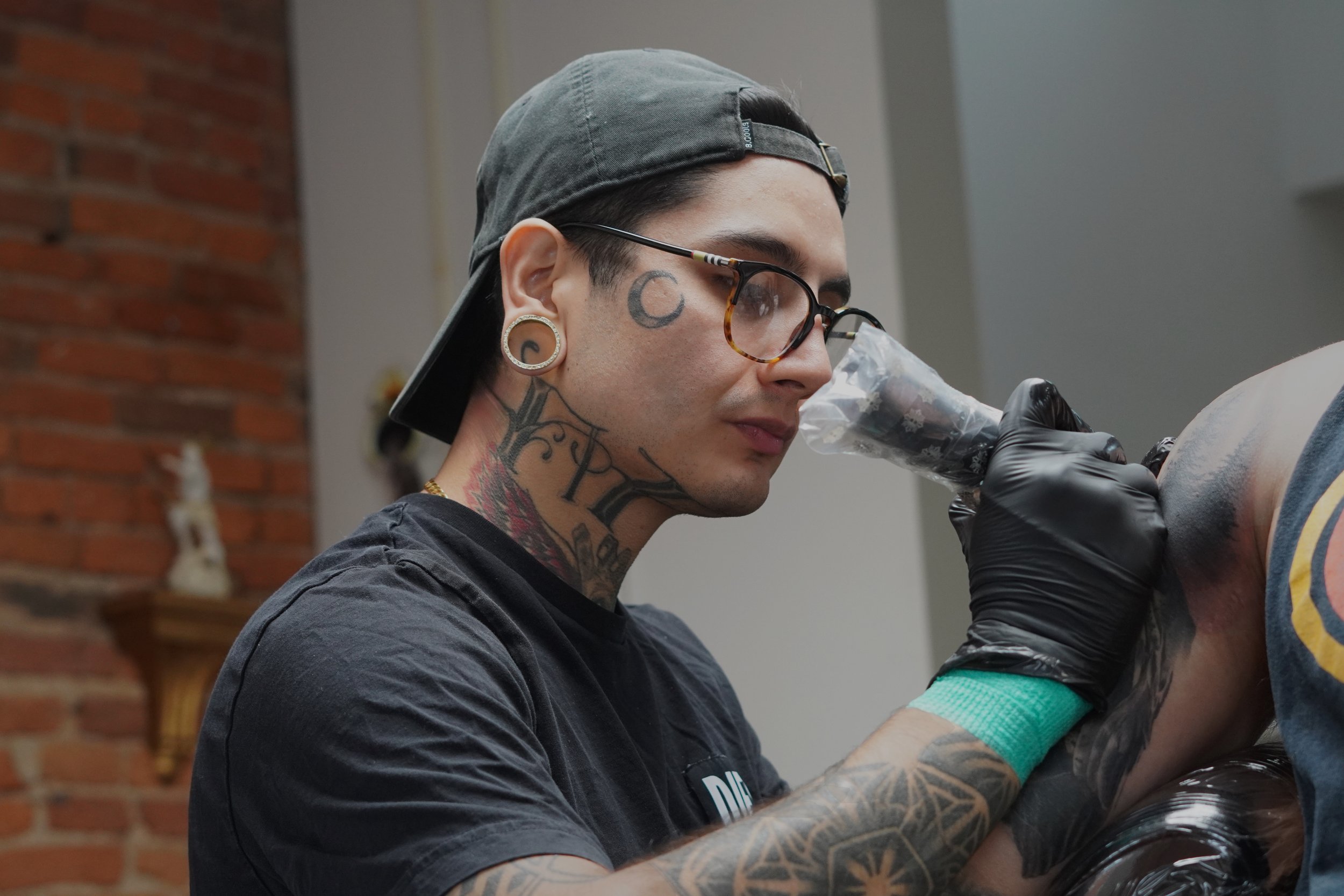 Tattoo artist wearing glasses, black gloves, and a cap, tattooing a person's shoulder in a tattoo studio. Cheap business photographer orange county ny northern nj