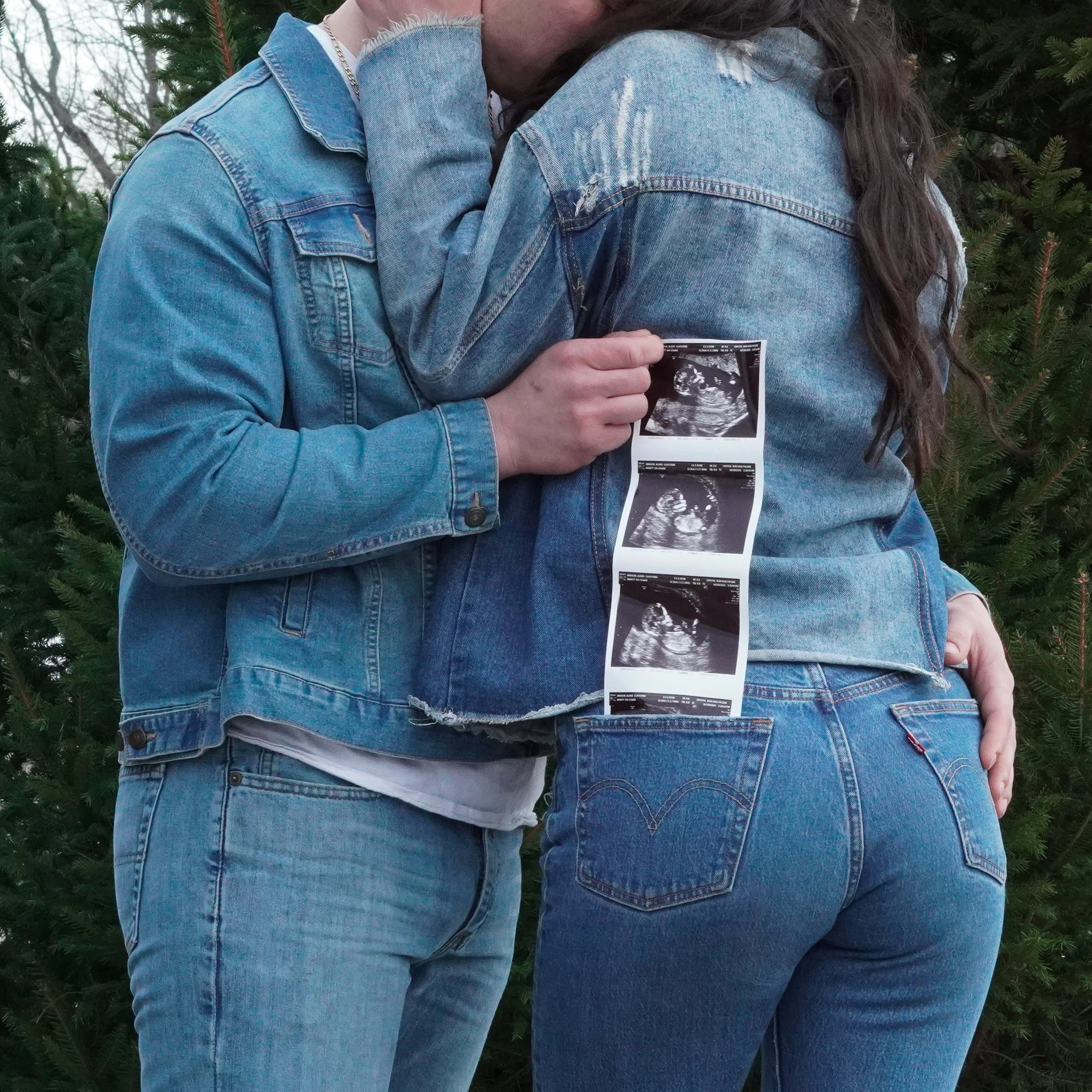 A couple standing outdoors in front of greenery, with the woman showing a series of ultrasound photos tucked into the back pocket of her jeans. Baby announcement photos.
