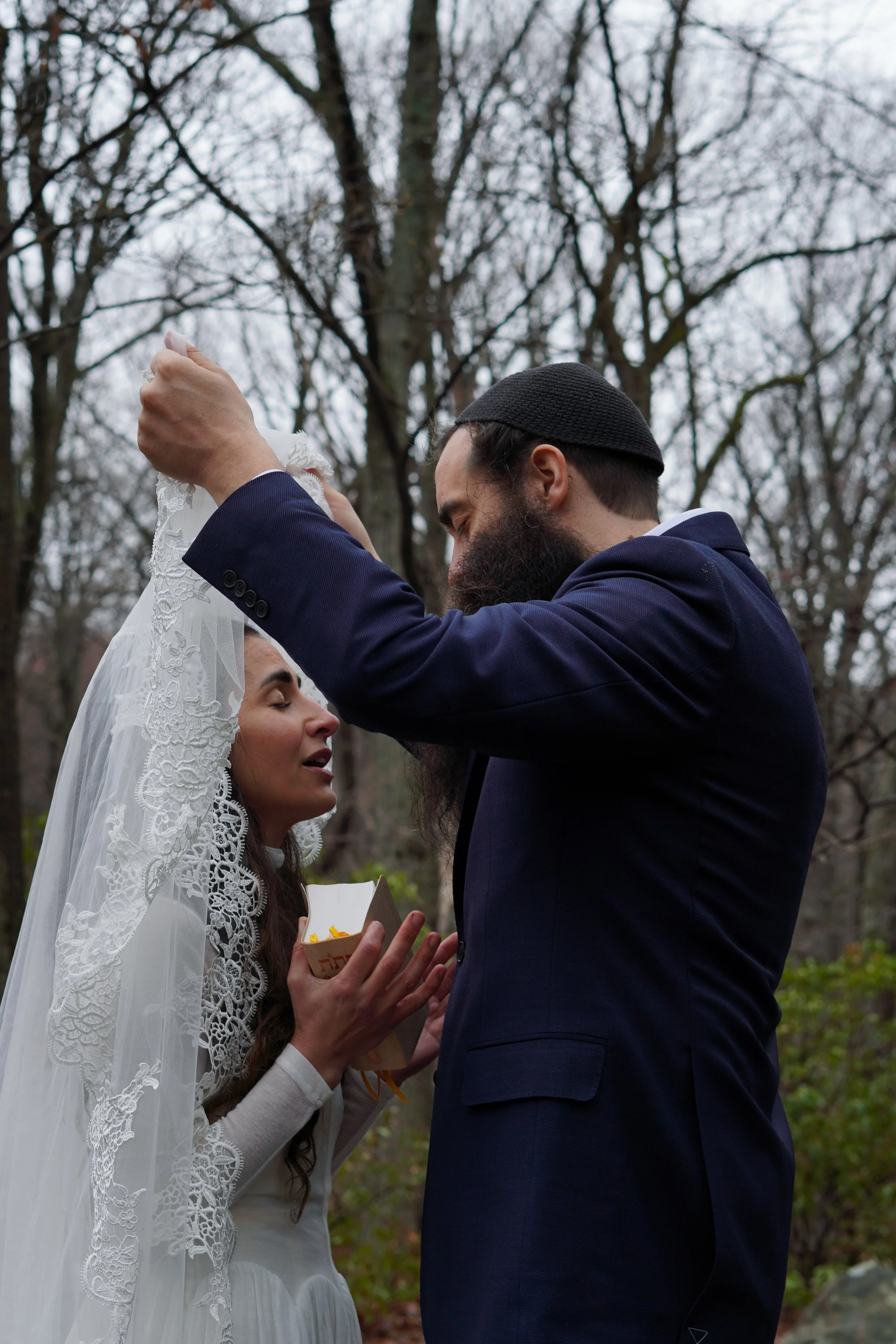 A couple exchanging wedding vows outdoors in a wooded area. The bride is holding a small box with yellow flowers, and the groom is placing a ring on her finger. The bride is wearing a white dress and lace veil, and the groom is wearing a dark suit an