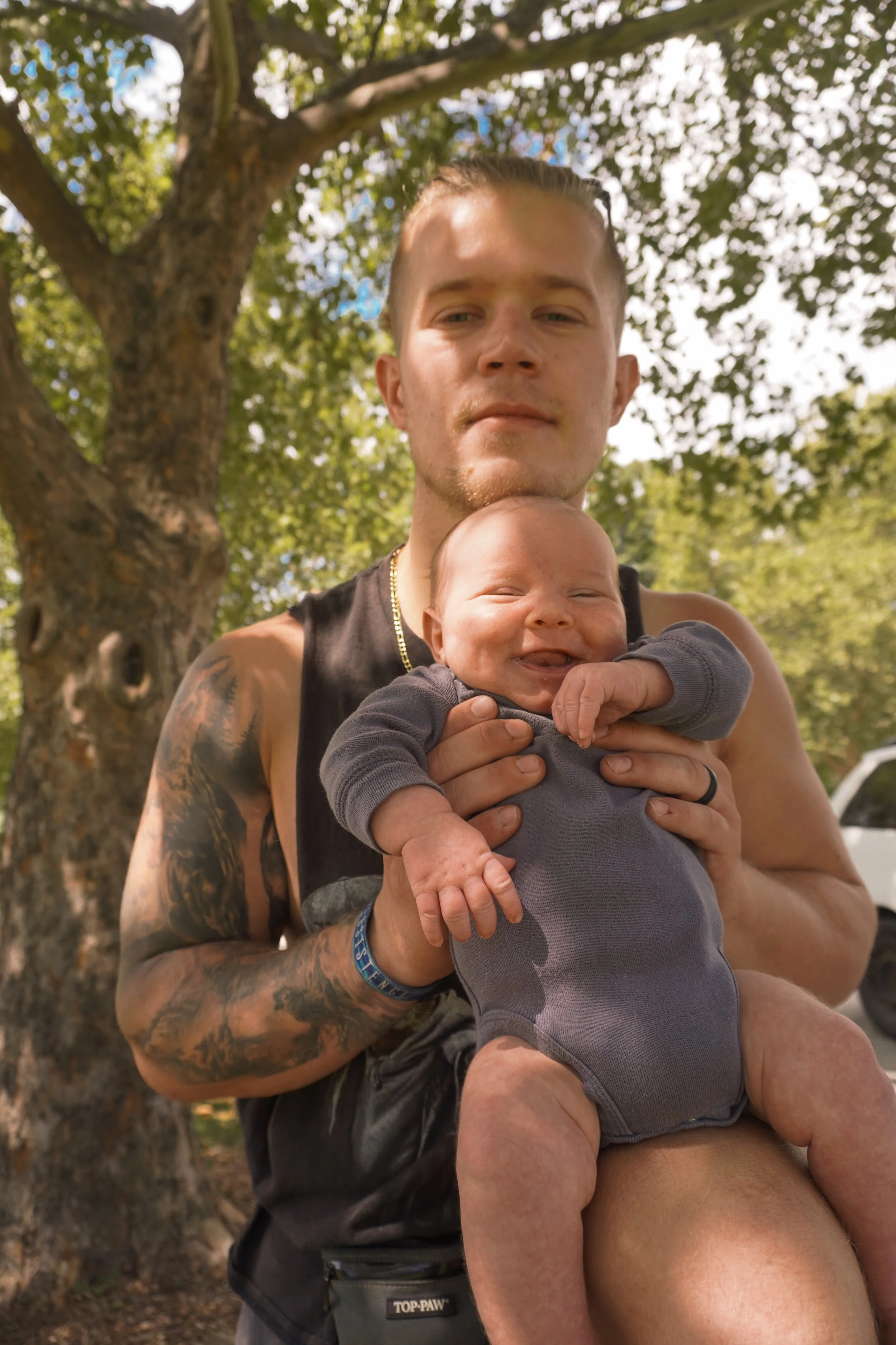 A young man with tattoos on his arm, holding a smiling baby, standing outdoors under a tree with green leaves. Family photos northern nj