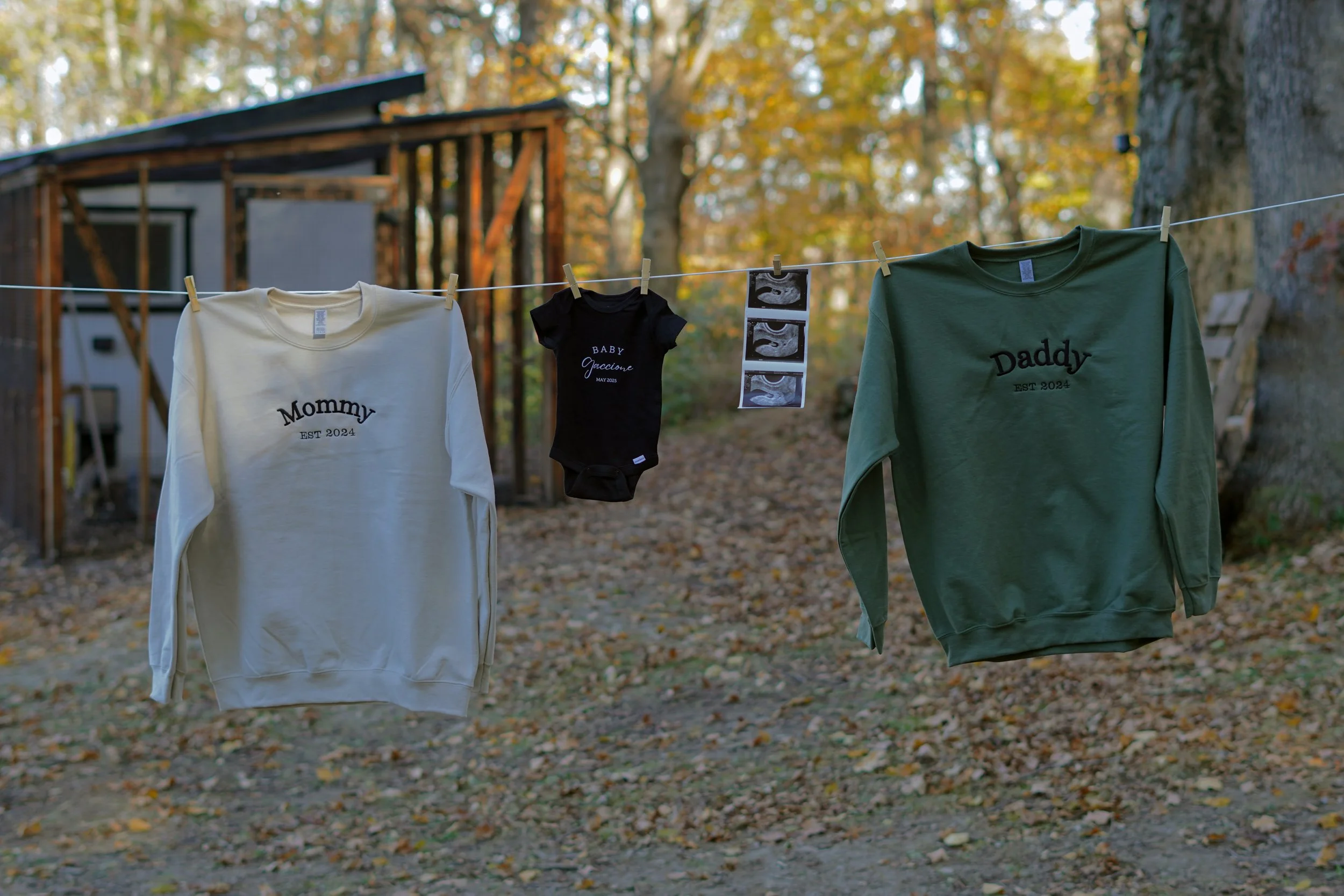 Clothes hanging on a clothesline outdoors with an autumn background; includes a white sweatshirt with 'Mommy EST 2024', a black baby onesie with 'Baby Jaccqueline May 2023', and a green sweatshirt with 'Daddy EST 2024'; ultrasound photos also hanging