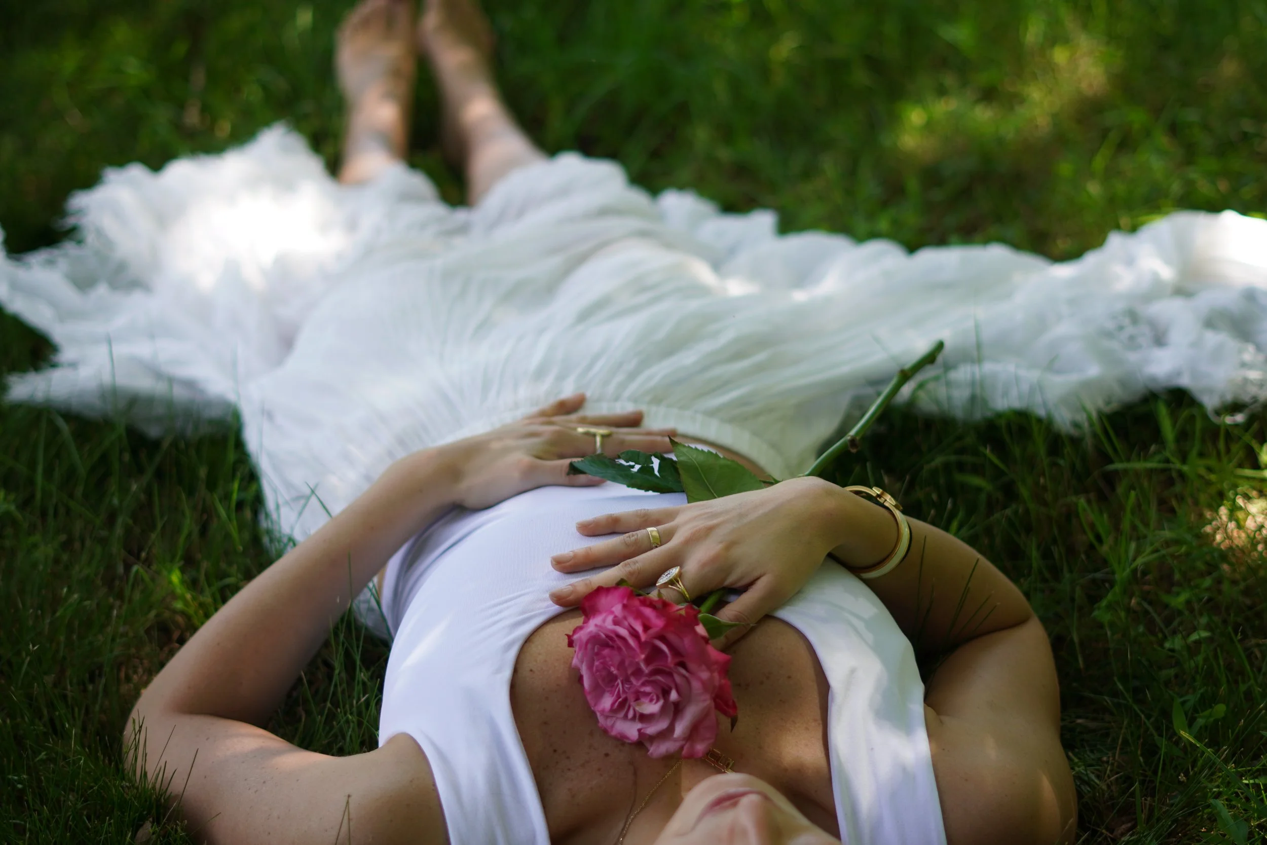 A woman lying on grass outdoors, wearing a white dress, holding a pink rose and green foliage, with jewelry on her hands and arm, in a relaxed pose.