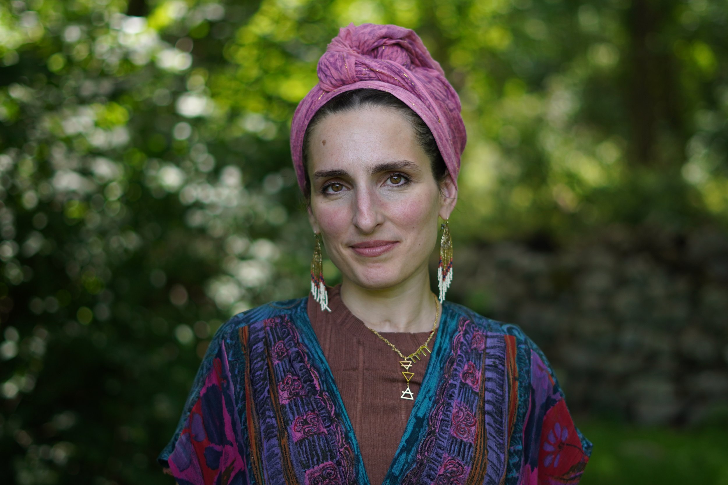 A woman with dark hair wearing a pink headscarf, earrings, and a colorful embroidered top standing outdoors with greenery in the background. Portrait photographer