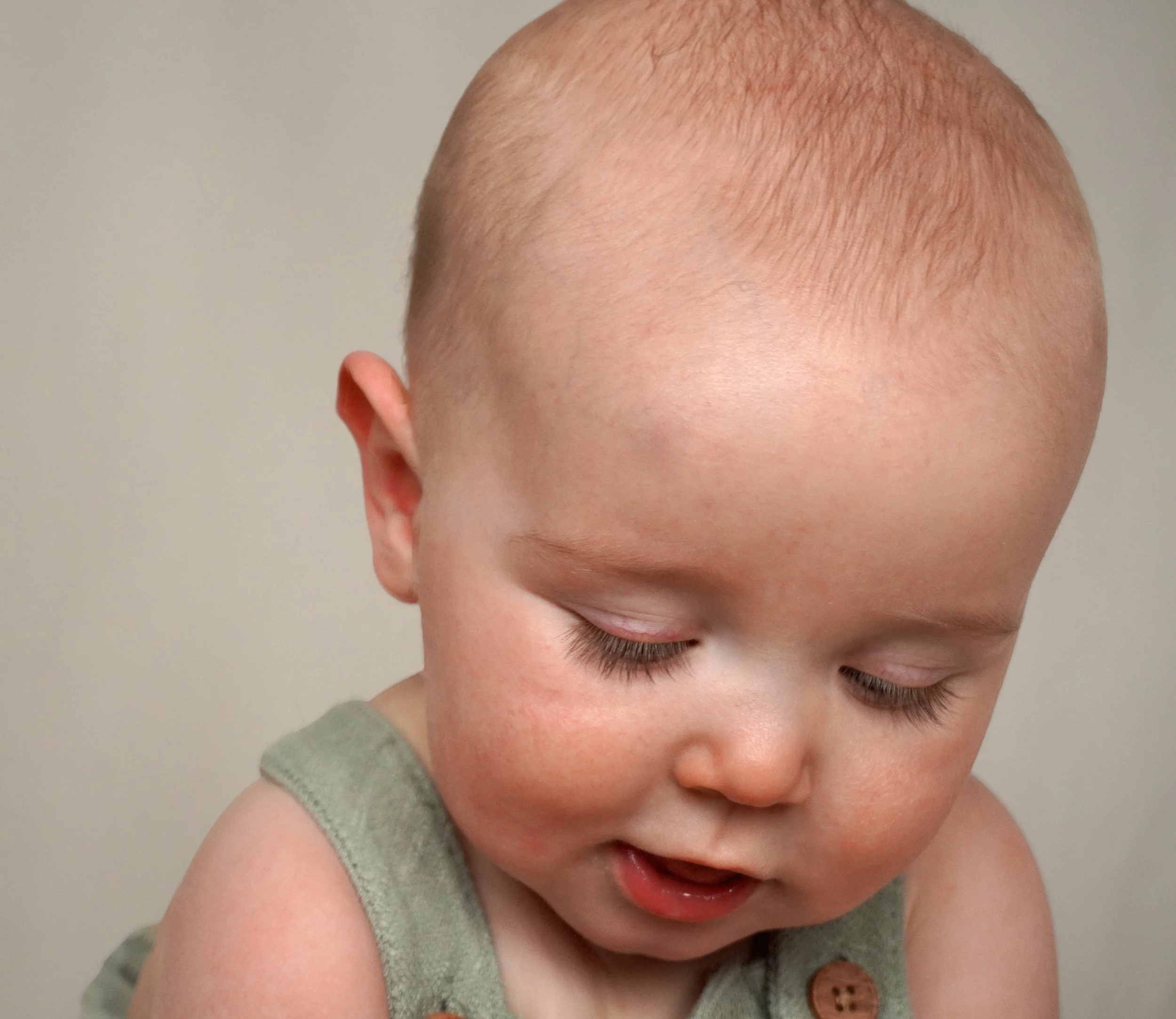 Close-up of a young child with light skin and a shaved head, wearing a green sleeveless shirt, looking downward with eyes closed. Baby photographer northern NJ