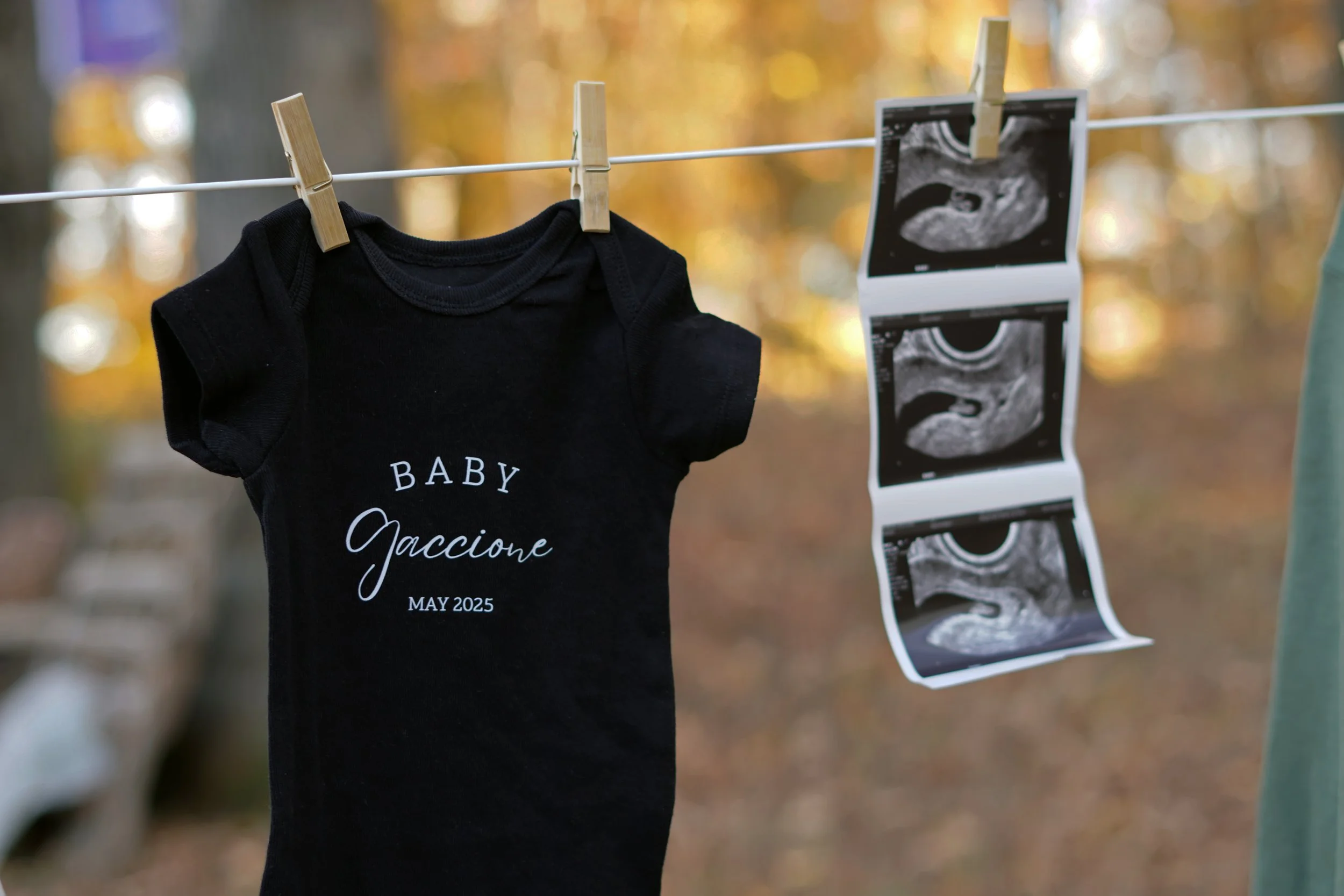 A black baby onesie hanging on a clothesline with the words 'Baby Gaccione May 2025' printed on it, next to ultrasound images also hanging on the line, in an outdoor setting with fall foliage. Baby announcement photographer northern nj