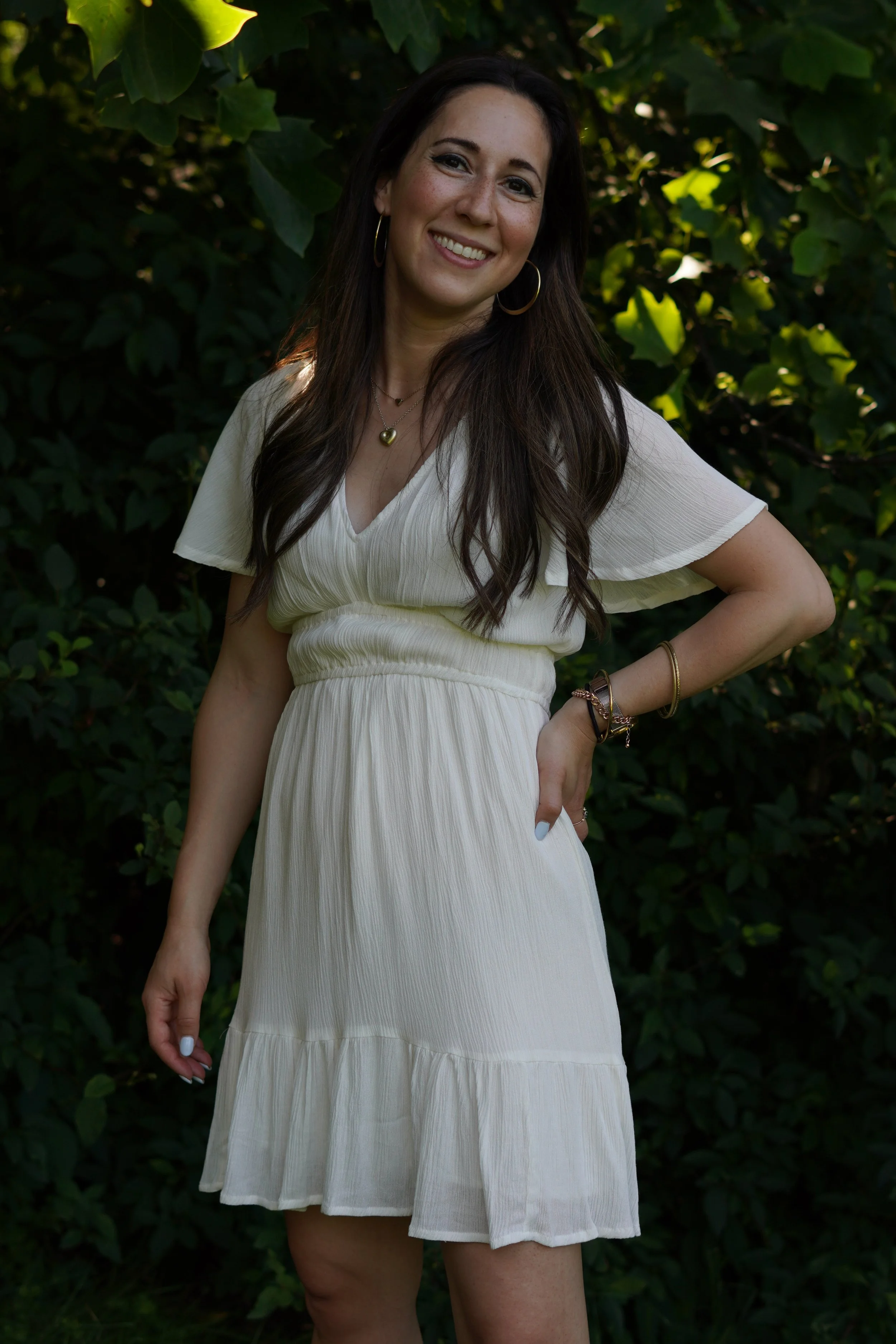 A woman with long dark hair and hoop earrings smiling outdoors in front of green foliage, wearing a flowing white dress with short sleeves, accessorized with necklaces and bracelets.