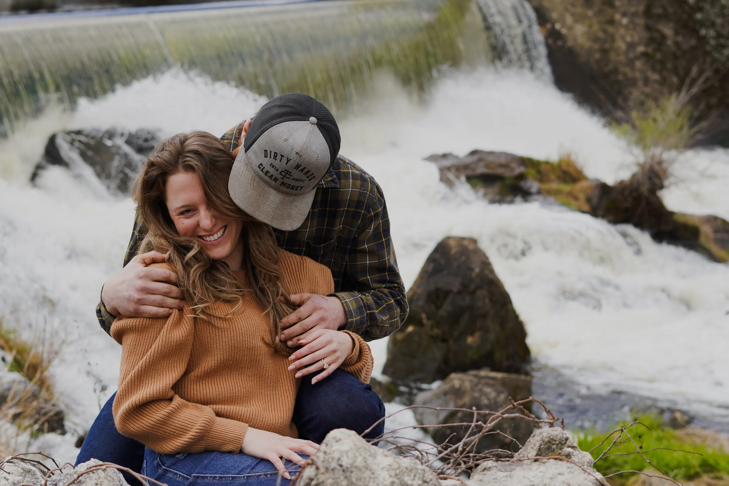 A young woman smiling and sitting on rocks near a flowing river or small waterfall, with a man hugging her from behind, in an outdoor natural setting. Cheap couple photographer northern nj