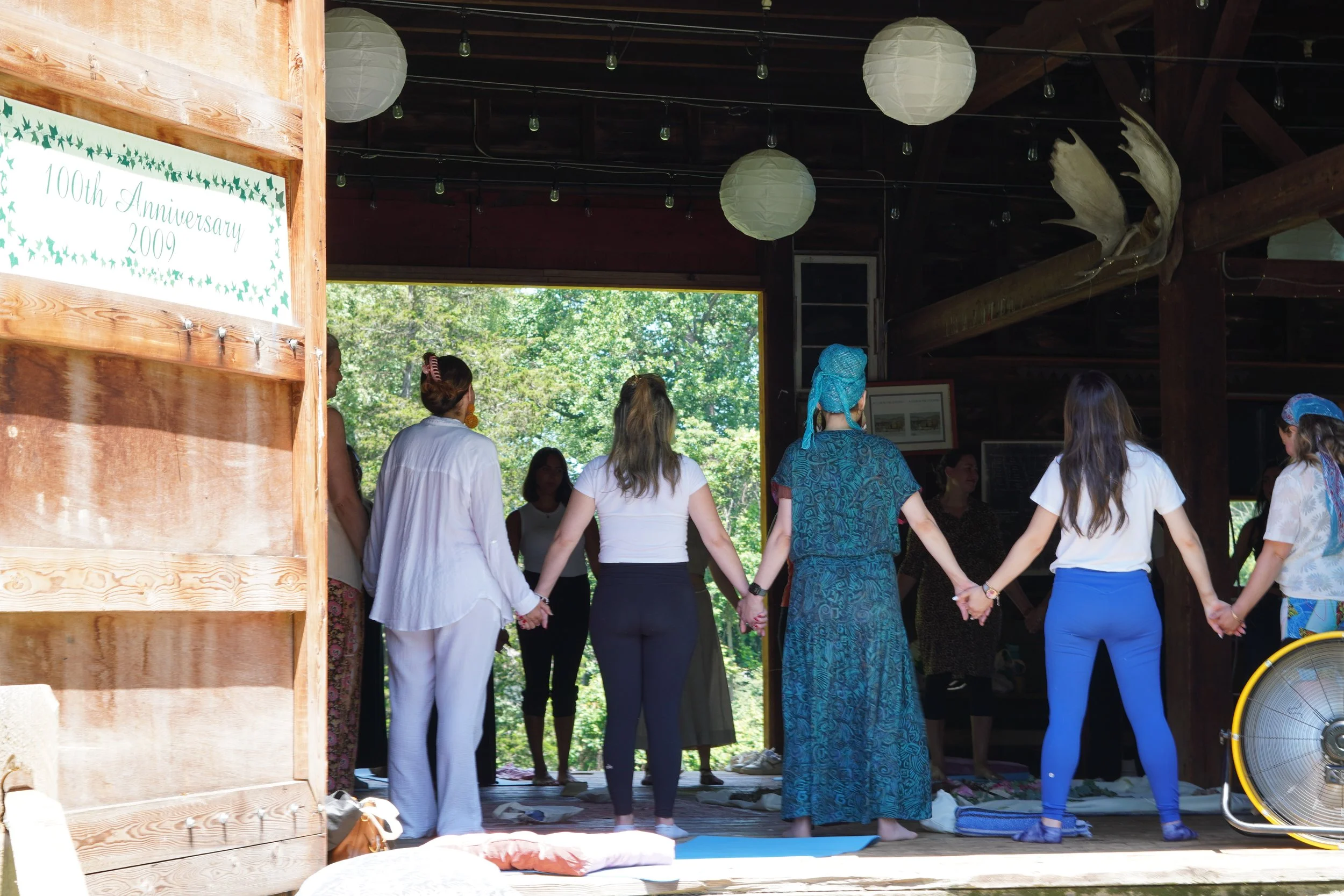 People holding hands in a circle during a gathering inside a wooden pavilion, with trees visible outside, and a sign on the left indicating a celebration of the 100th anniversary in 2009.