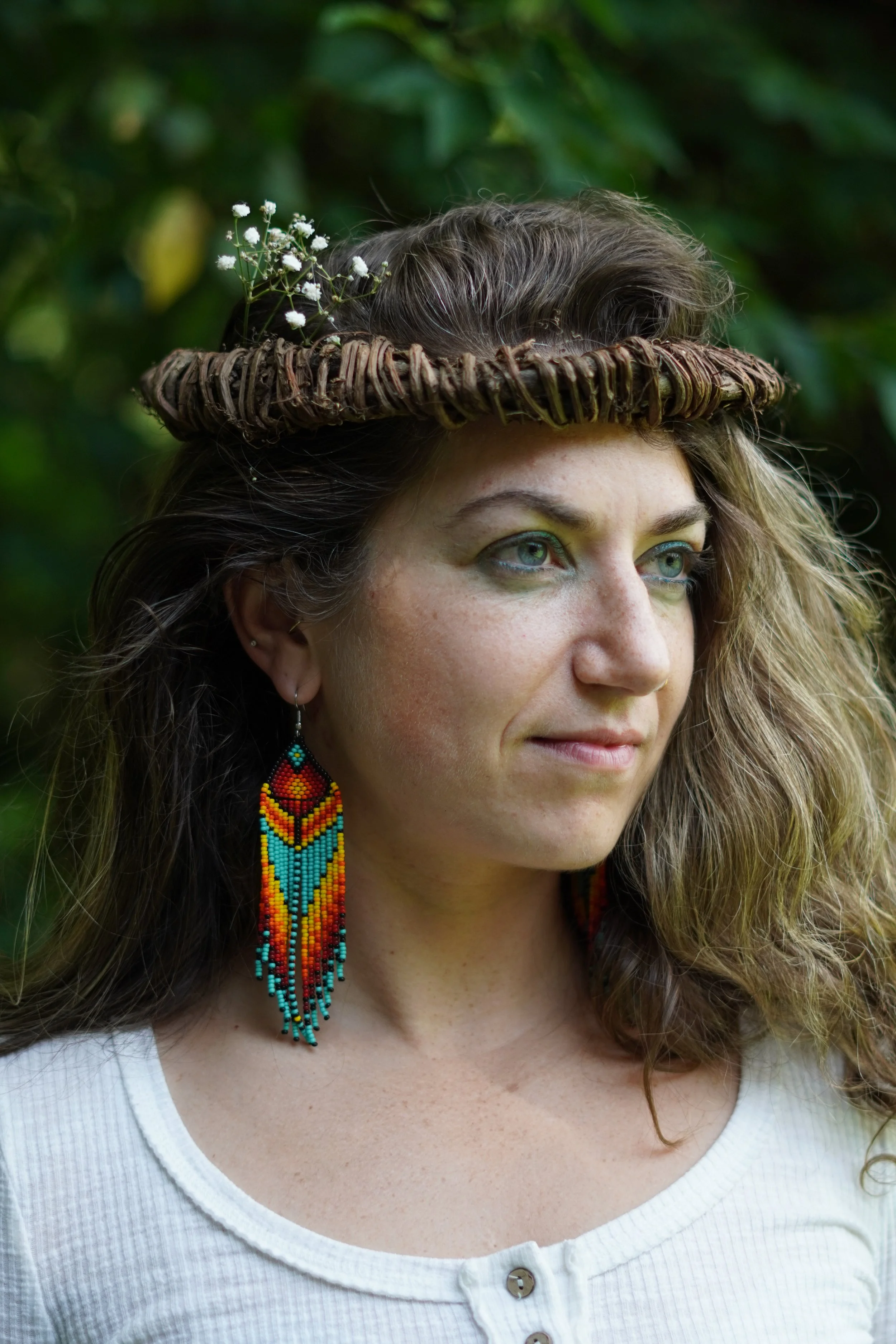A woman with long, curly hair wearing a woven headband, colorful beaded earrings, and a white top, outdoors with green foliage in the background. Portrait photographer