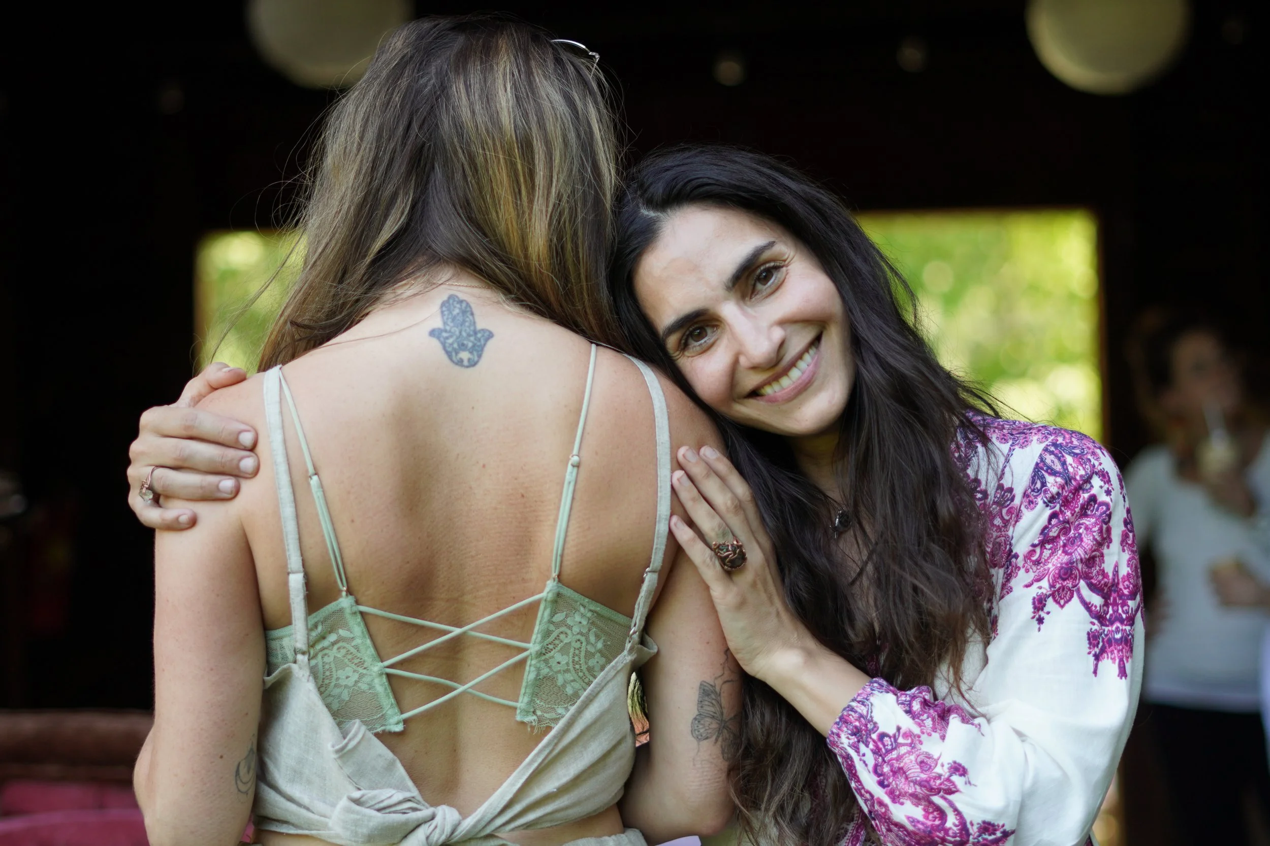 Two women hugging, with one woman smiling at the camera. The woman facing away has a tattoo of a blue charm on her shoulder and is wearing a beige top with lace details and crisscross back straps. The woman smiling has long dark hair and is wearing a