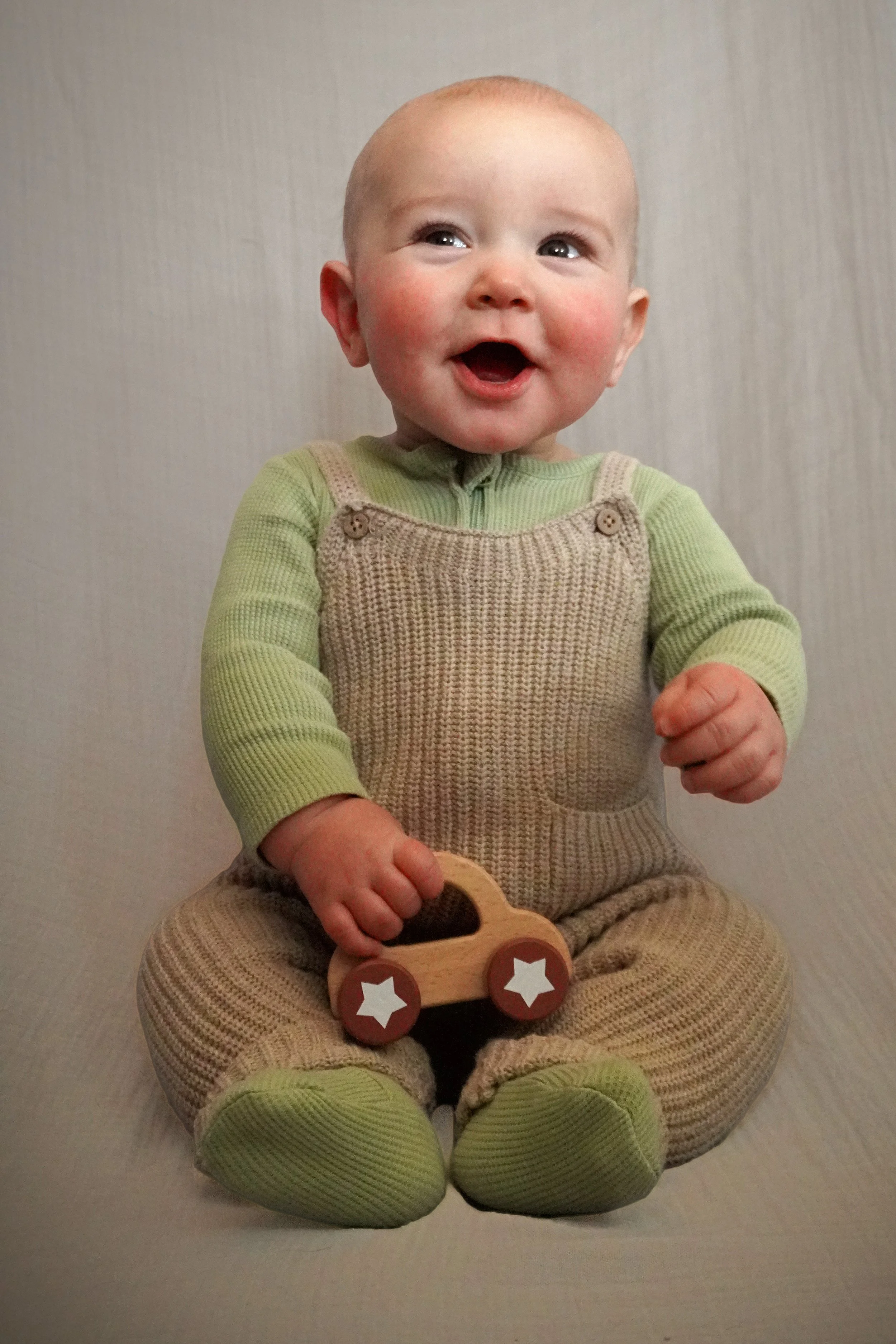 A smiling baby sitting on the floor, holding a wooden toy car, dressed in a green and beige knitted outfit. Baby photography northern nj