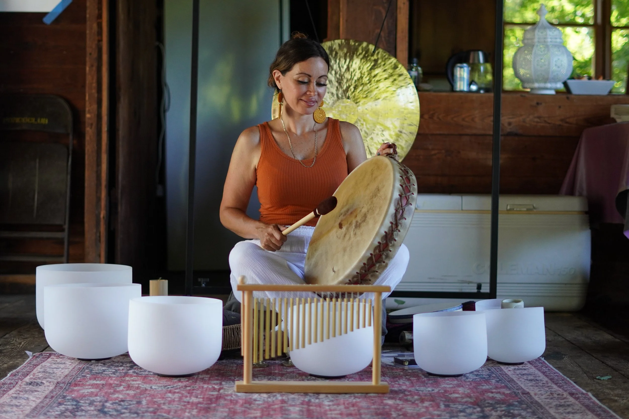 Woman playing crystal bowls and drum in a cozy wooden room with large window, potted plant, and decorative items