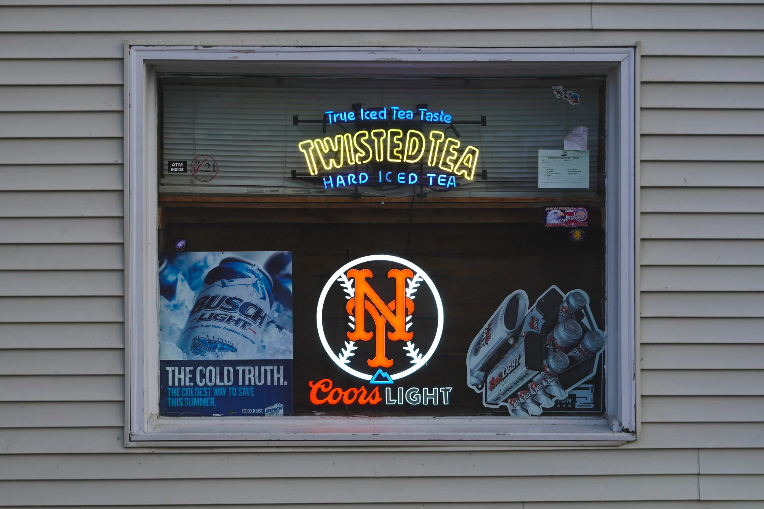Neon sign advertising twisted iced tea, hard iced tea, and true iced tea taste in a store window with a Bud Light poster, a Coors Light logo, and a six-pack of beer cans. Retro photographer cheap northern nj