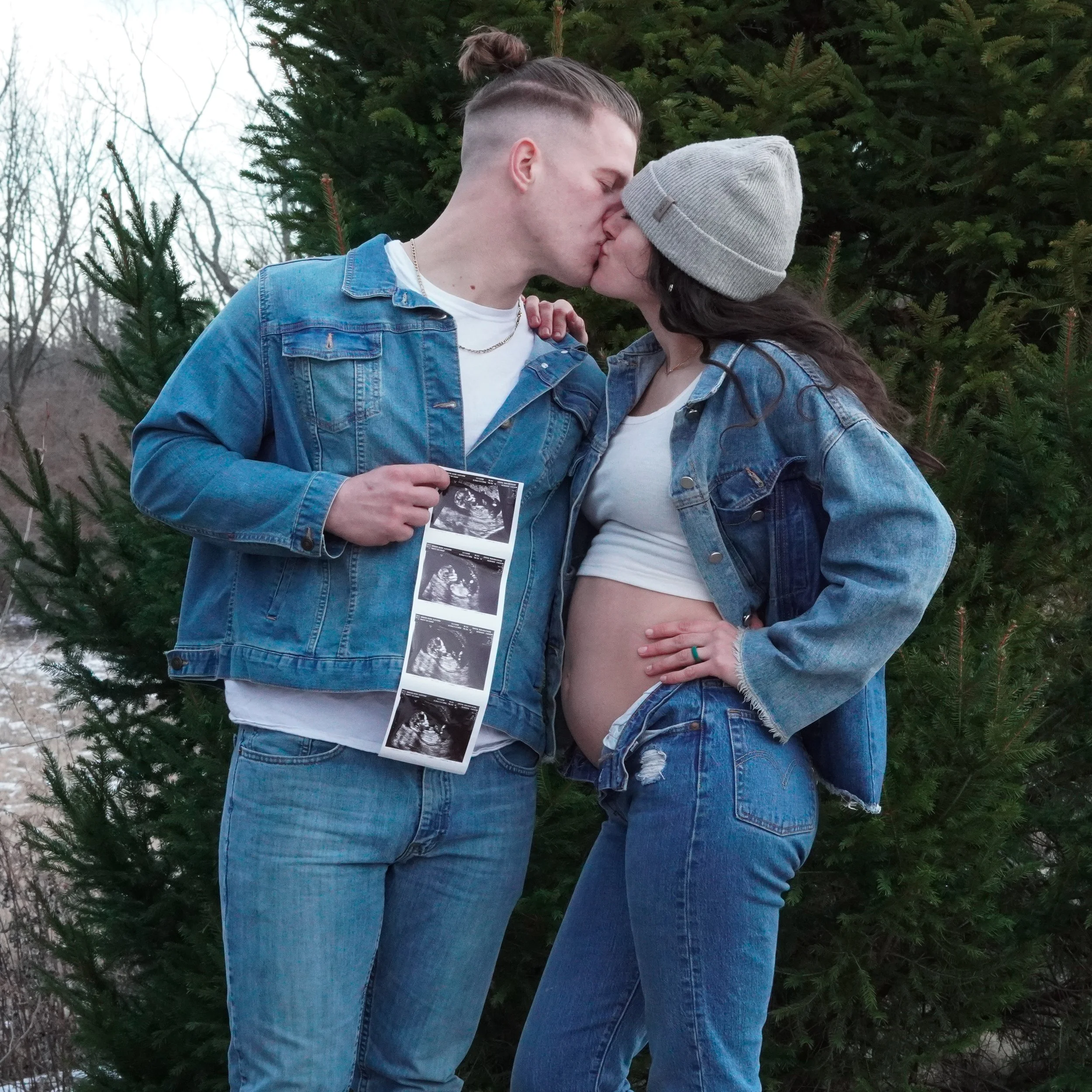 A couple kissing outdoors with snow and trees in the background. The woman is pregnant, wearing a beanie and denim jacket, and the man is holding ultrasound pictures. Baby announcement photos