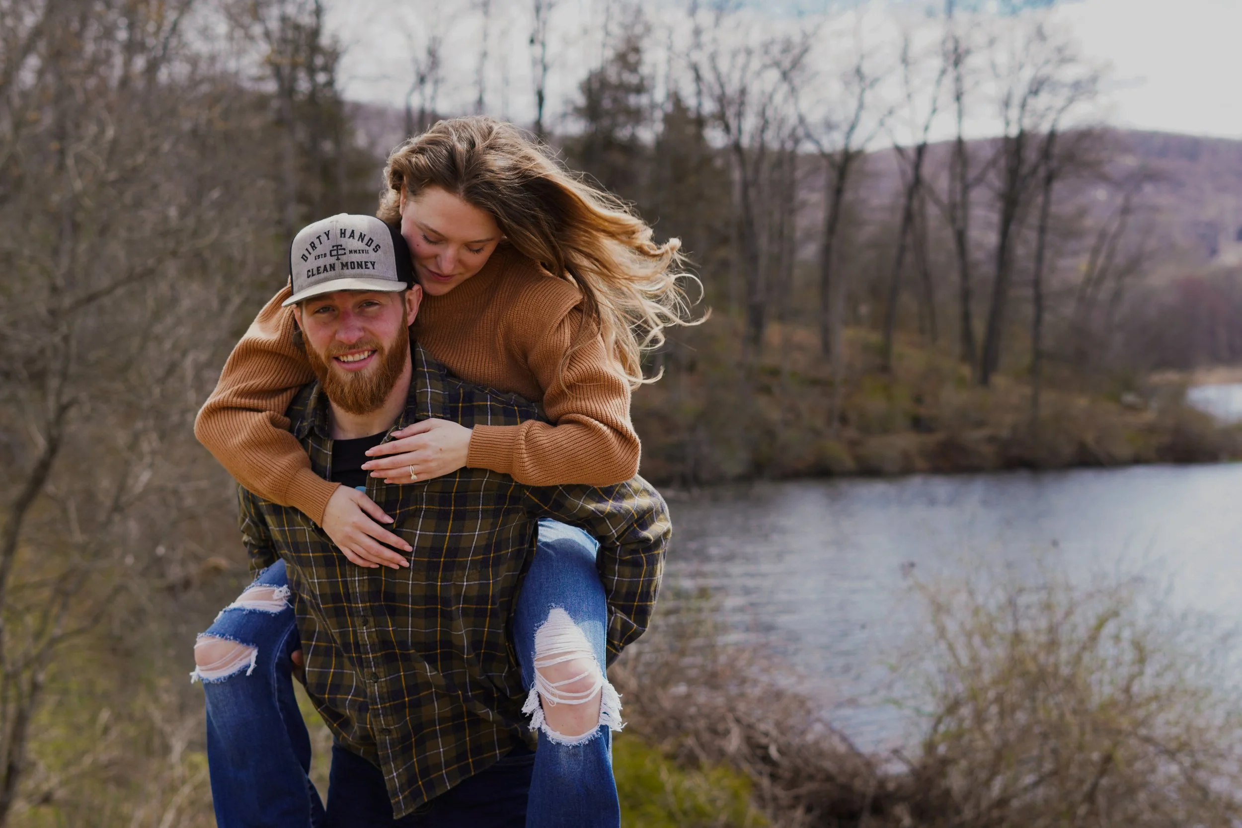 A young couple outdoors near a river, with trees in the background. The woman is piggybacking on the man, both smiling and happy.