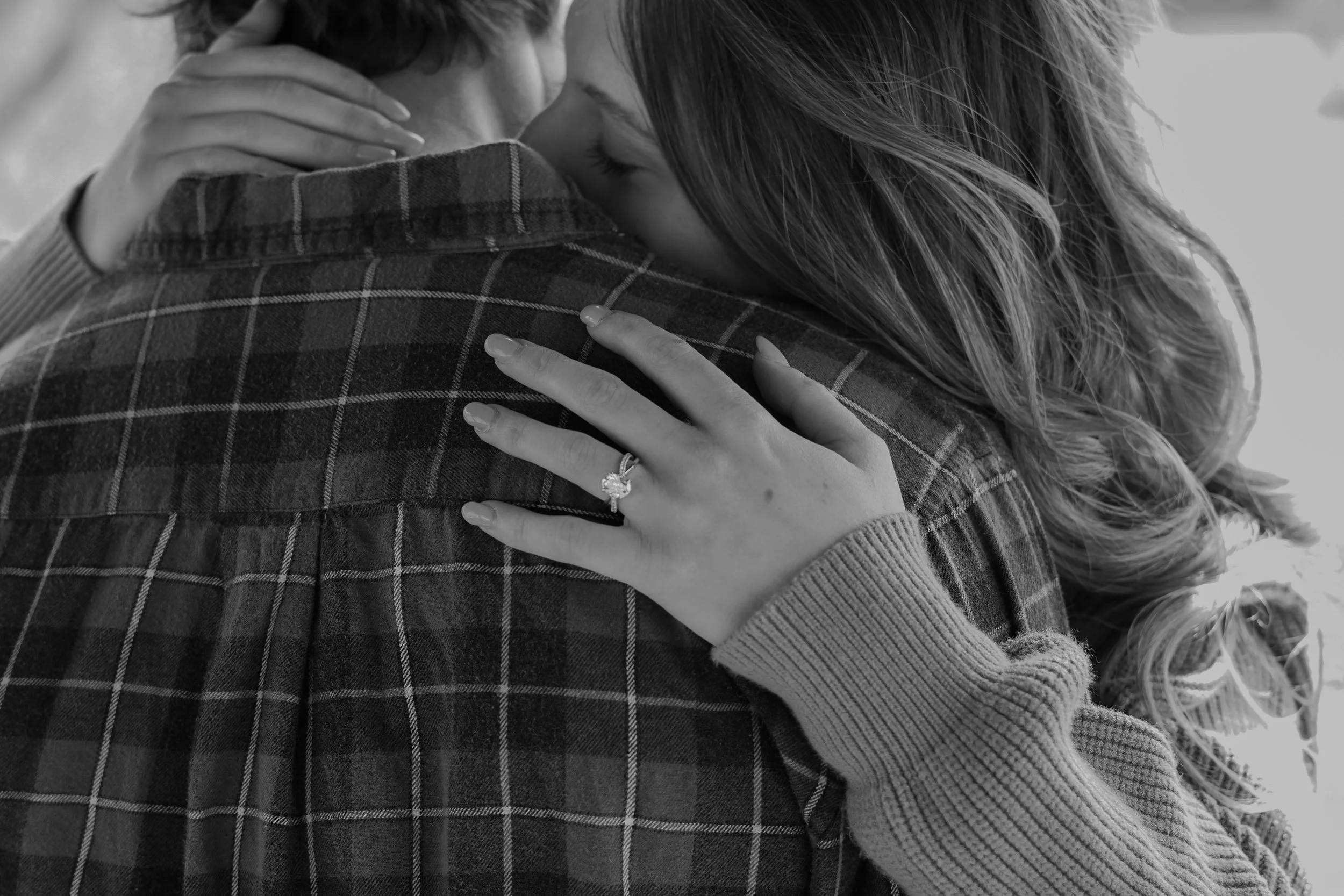 A woman with wavy hair is hugging a man wearing a checkered shirt. The woman's left hand with a ring on her ring finger is resting on the man's shoulder.