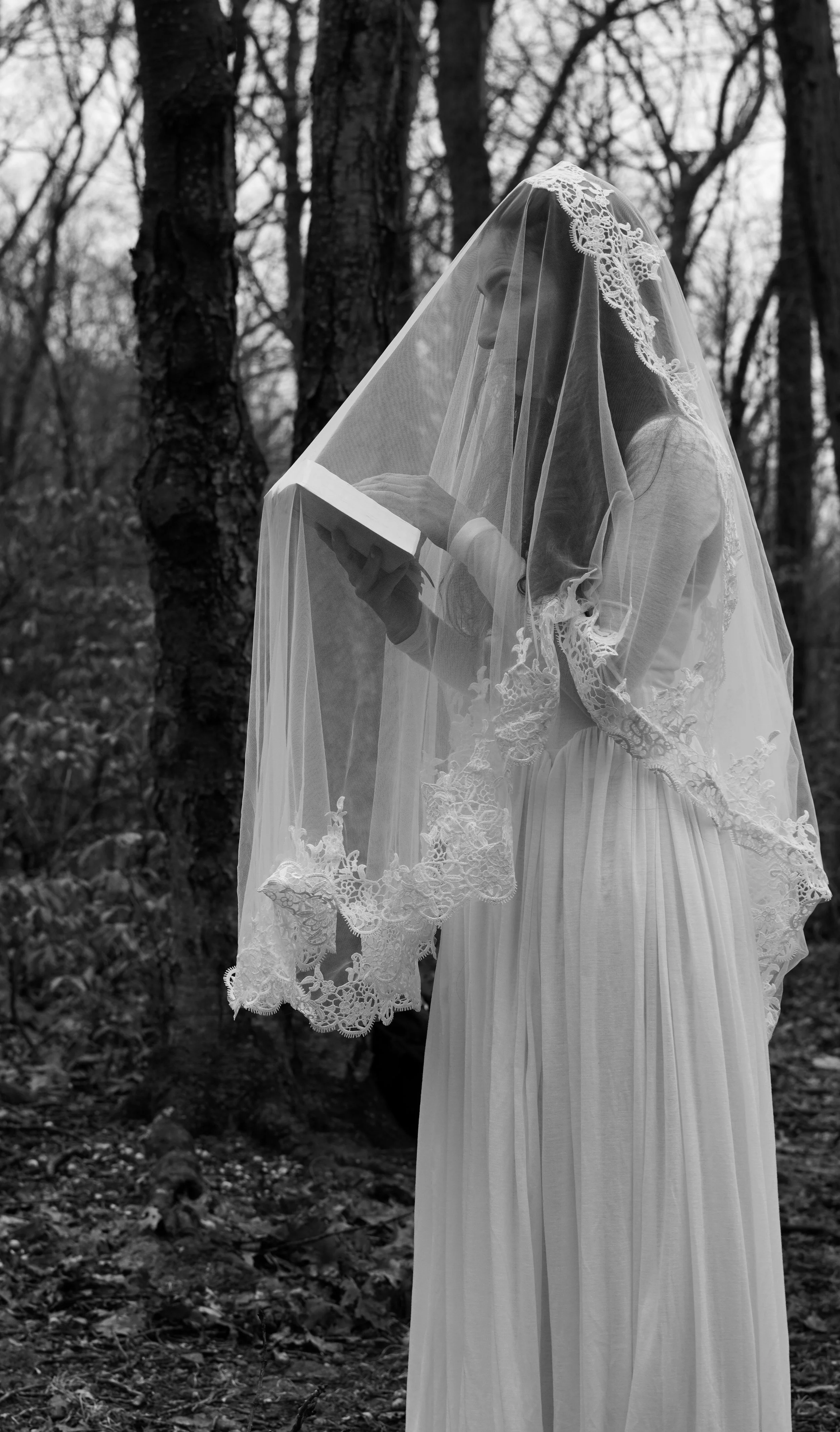 A woman in a long white dress and lace veil holds a book, standing outdoors in a wooded area, captured in black and white. Wedding photographer northern nj