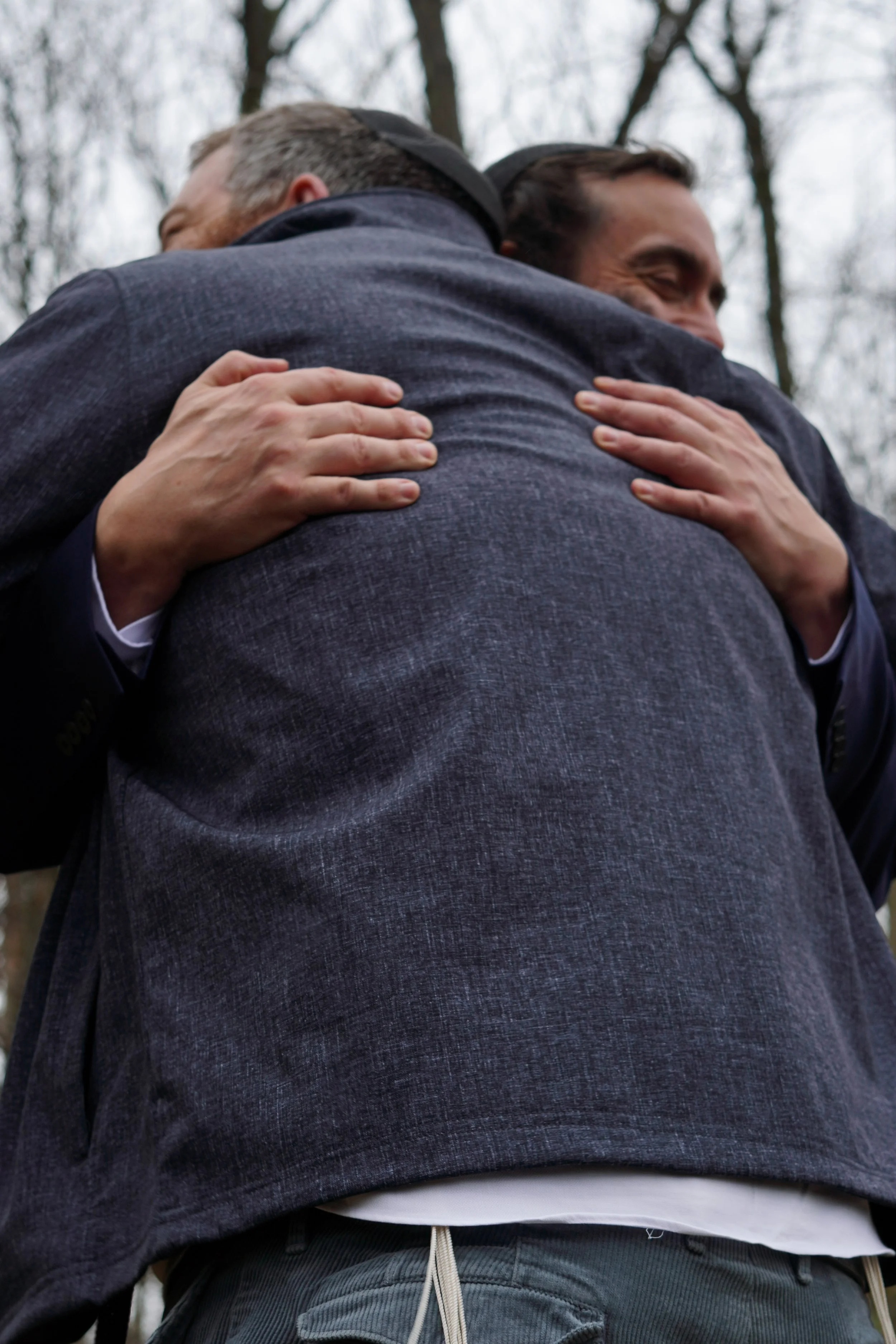 Two men hugging outdoors, in a forested area during daytime, sharing a warm embrace. Cheap wedding photographer orange county ny