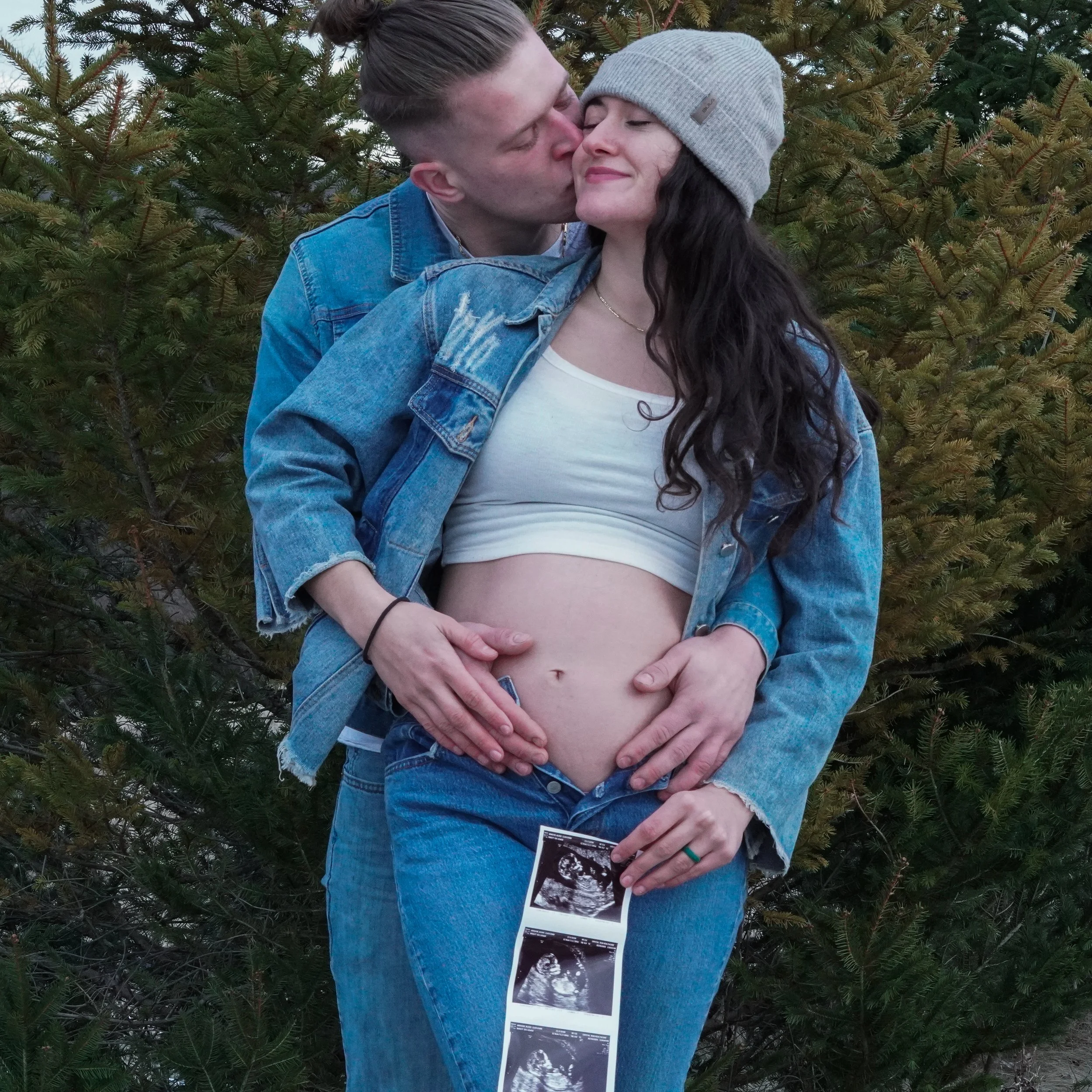 A couple standing outdoors in front of evergreen trees; the woman is pregnant, and the man is kissing her cheek. She is holding ultrasound images that show a developing fetus. Baby announcement photos