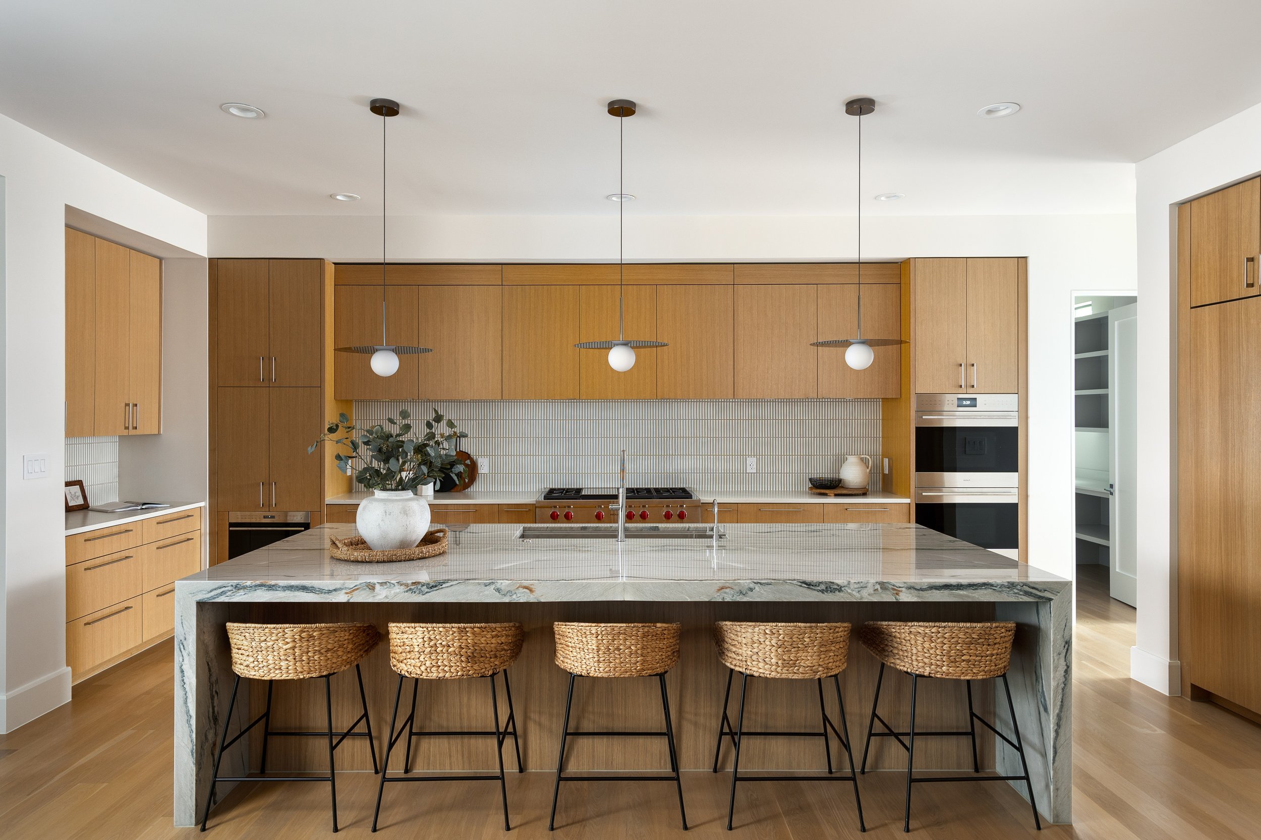 Modern kitchen with wooden cabinets, a large marble island with woven stools, pendant lighting, and a neutral color palette.