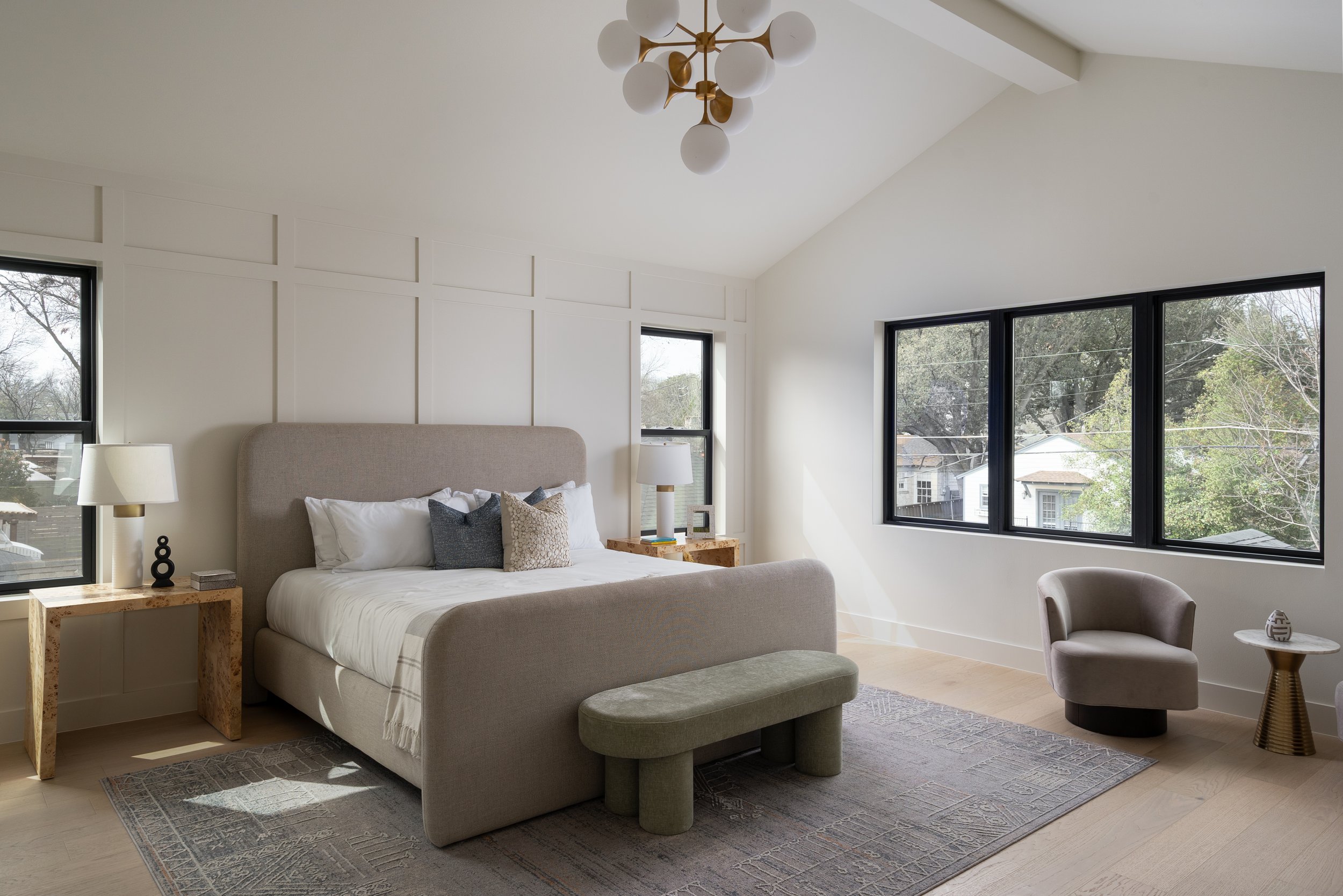 A modern bedroom with a beige upholstered bed, decorative pillows, and nightstands with lamps. Large windows with black frames let in natural light, showing trees and residential houses outside. The ceiling has a white geometric light fixture.