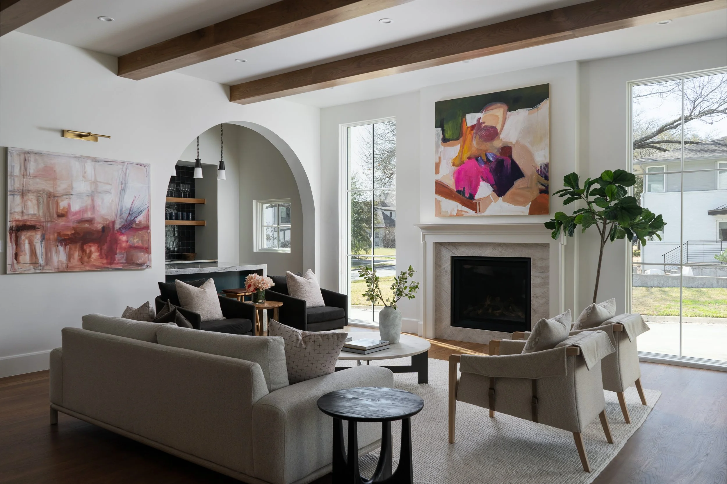 Living room with white walls, large windows, a fireplace, and modern art paintings on the wall. There are multiple couches and chairs arranged around a coffee table with flowers, with natural light coming in through the windows.