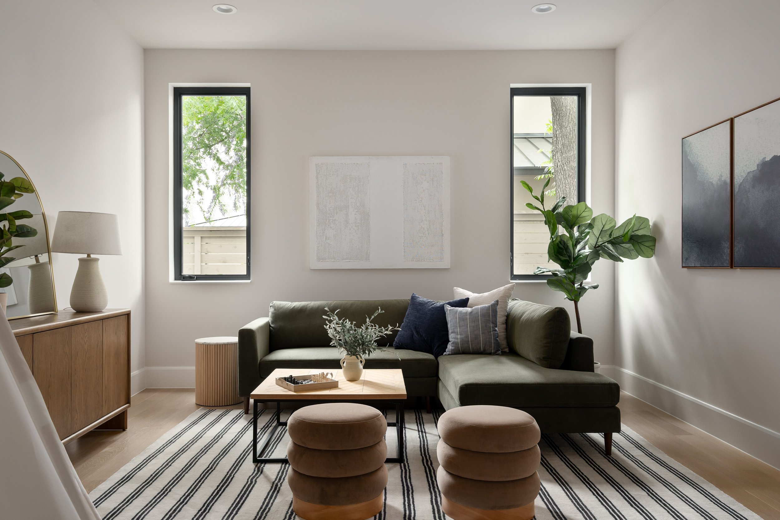 A living room with two tall windows on the back wall letting in natural light and showing green trees outside. It has a dark green sofa with throw pillows, a wooden coffee table with a vase and small decorative items, four brown rounded ottomans, a b