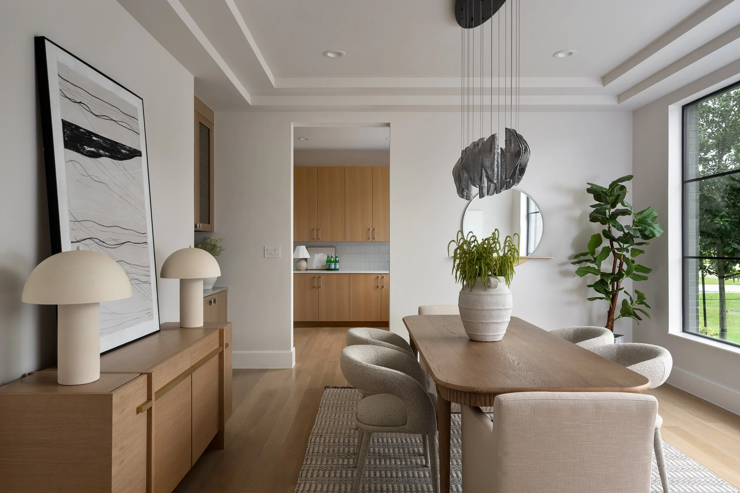 Modern interior living room and dining area with wood furniture, green plants, wall art, and large windows overlooking greenery.