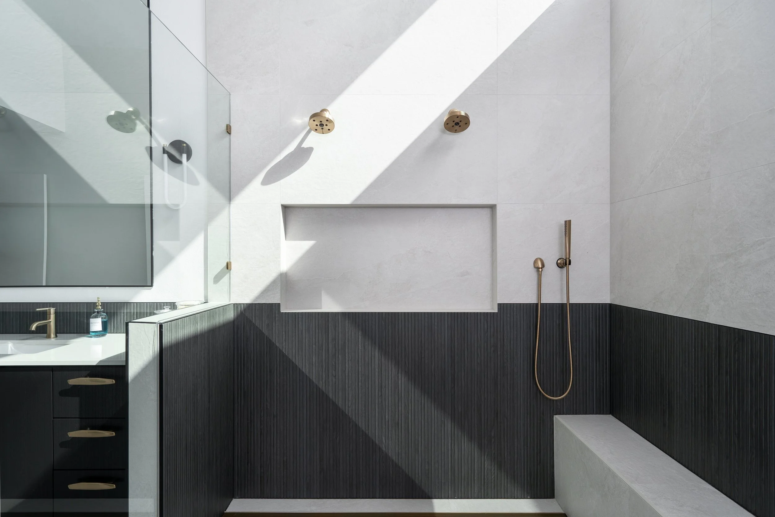 Modern bathroom with white walls, black cabinetry, a large mirror, and a sunlit shower area with brass fixtures and a built-in bench.