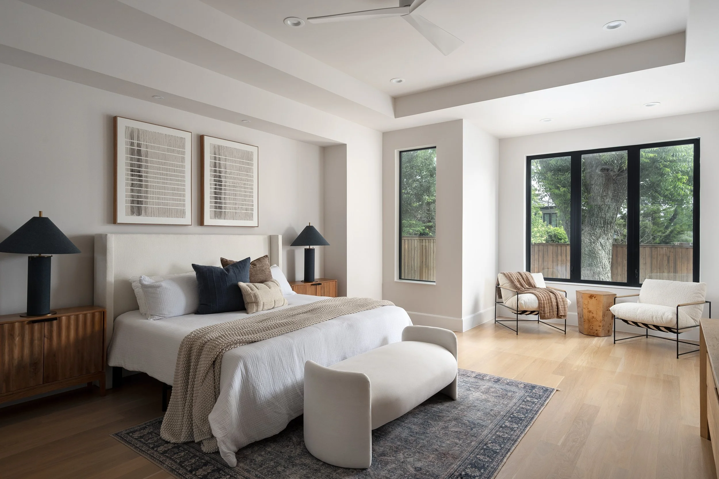 A modern living room with white walls, large windows, a white upholstered sofa, a wooden side table, black lamps, framed artwork, and additional seating with blanket accents.