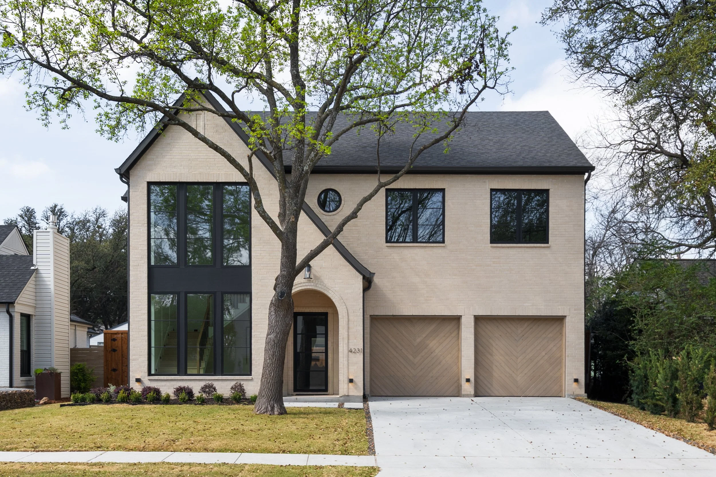 A modern two-story house with beige brick exterior, large black framed windows, and a black shingled roof. There is a large tree in front with green leaves and a small lawn with some plants.