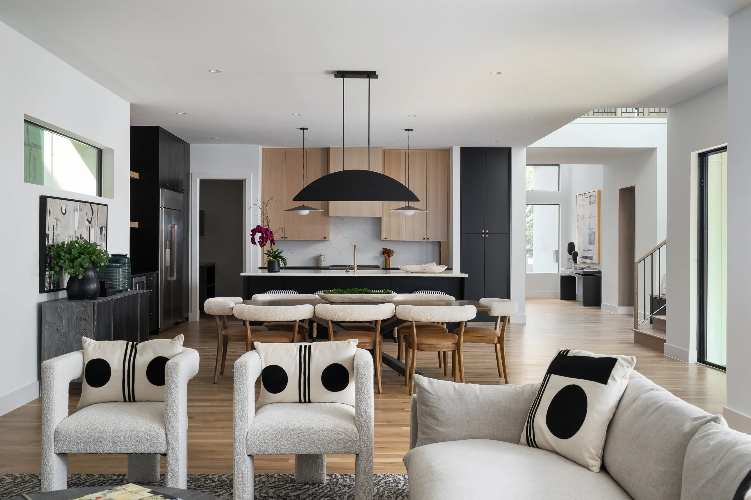 Modern open-concept living and dining area with black, white, and natural wood decor; white armchairs with black polka dots in the foreground, a dining table with wooden chairs in the middle, and a kitchen with wooden cabinets and black accents in th