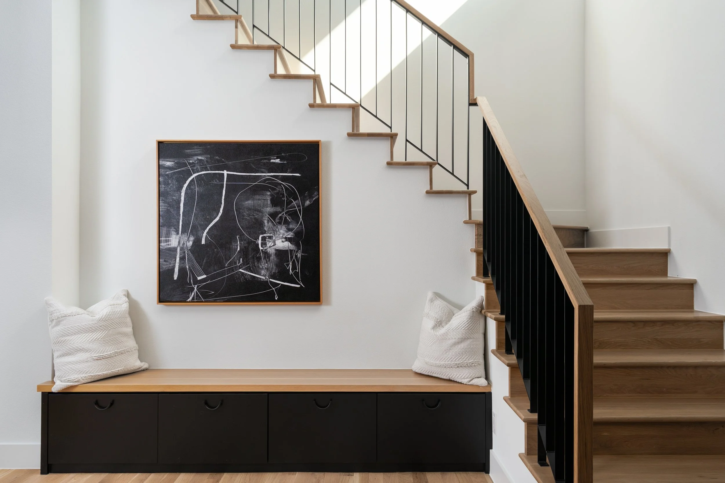 Interior of a modern home staircase with wooden steps and black metal railing. A black cabinet with three drawers and a wooden top is underneath, decorated with two beige textured pillows. A large abstract black and white artwork hangs on the wall ab