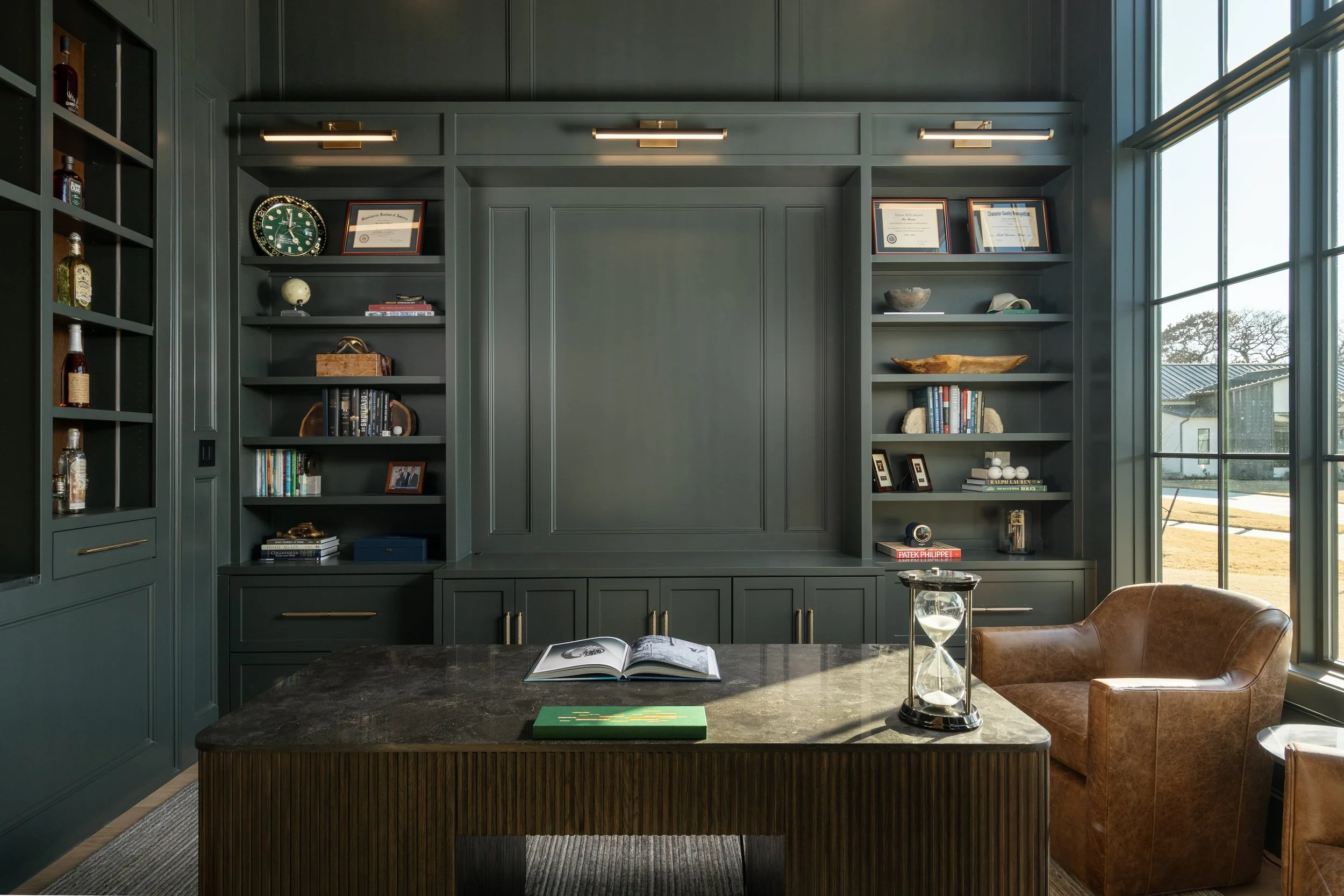 Interior of a room with dark green built-in bookshelves and cabinets, a large window on the right, a marble top table with an open book and a glass hourglass, a brown leather chair, and various decorative items and framed certificates on the shelves.