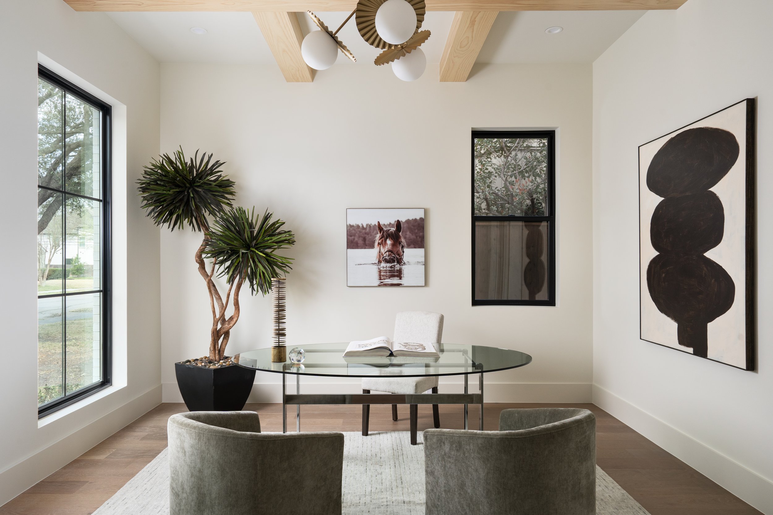 Modern dining room with a round glass table, four upholstered chairs, a large potted plant, framed animal photography, abstract artwork, and a window bringing in natural light.