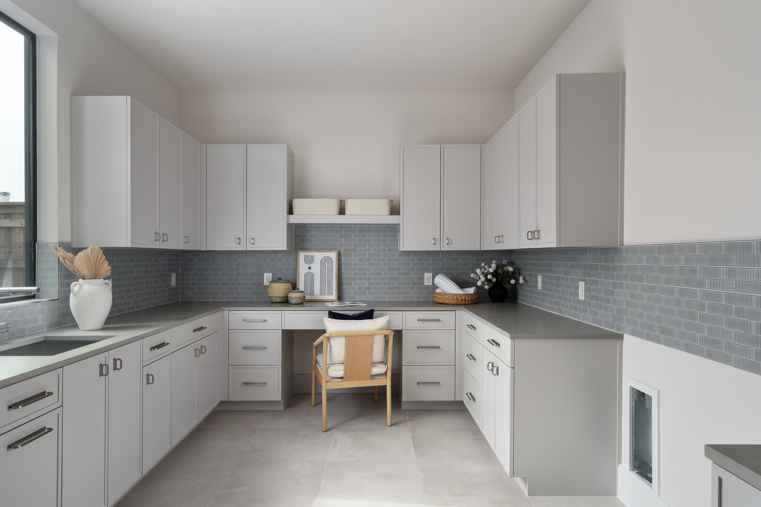 Modern kitchen with white cabinets, a gray tiled backsplash, and a gray countertop. Decor includes a white vase with dried plants, framed artwork, small baskets, and a vase with flowers. A wooden chair with cushions is in the center, under a desk-lik