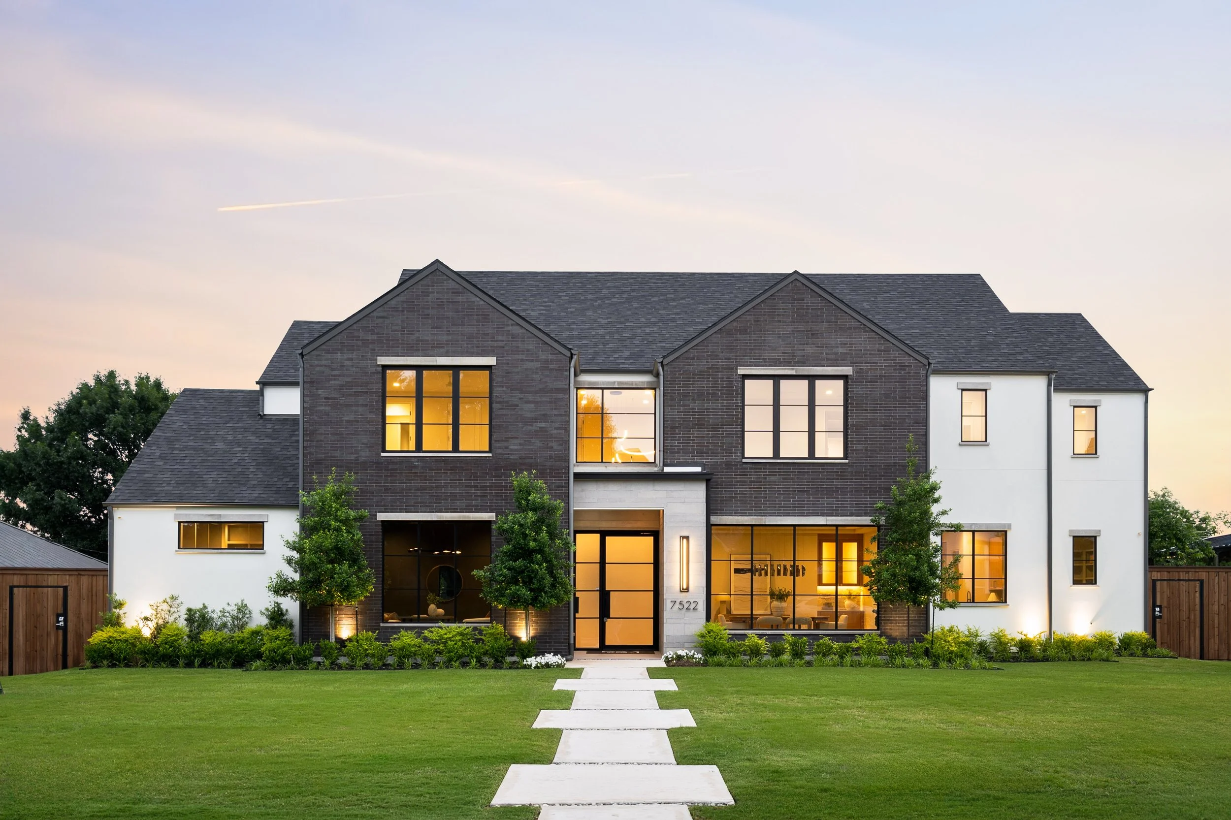 Modern two-story house with a dark brick facade and large illuminated windows, surrounded by a manicured lawn and small trees, under a clear sky at dusk.