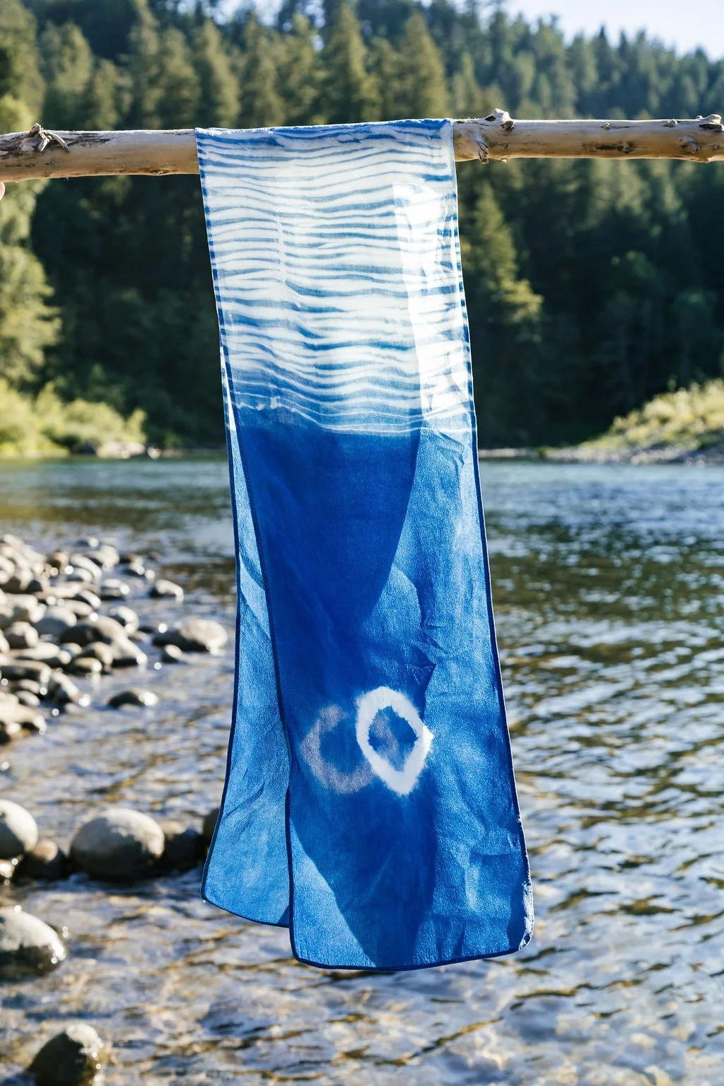 A blue tie-dye cloth hanging on a wooden stick over a river with a forested background.