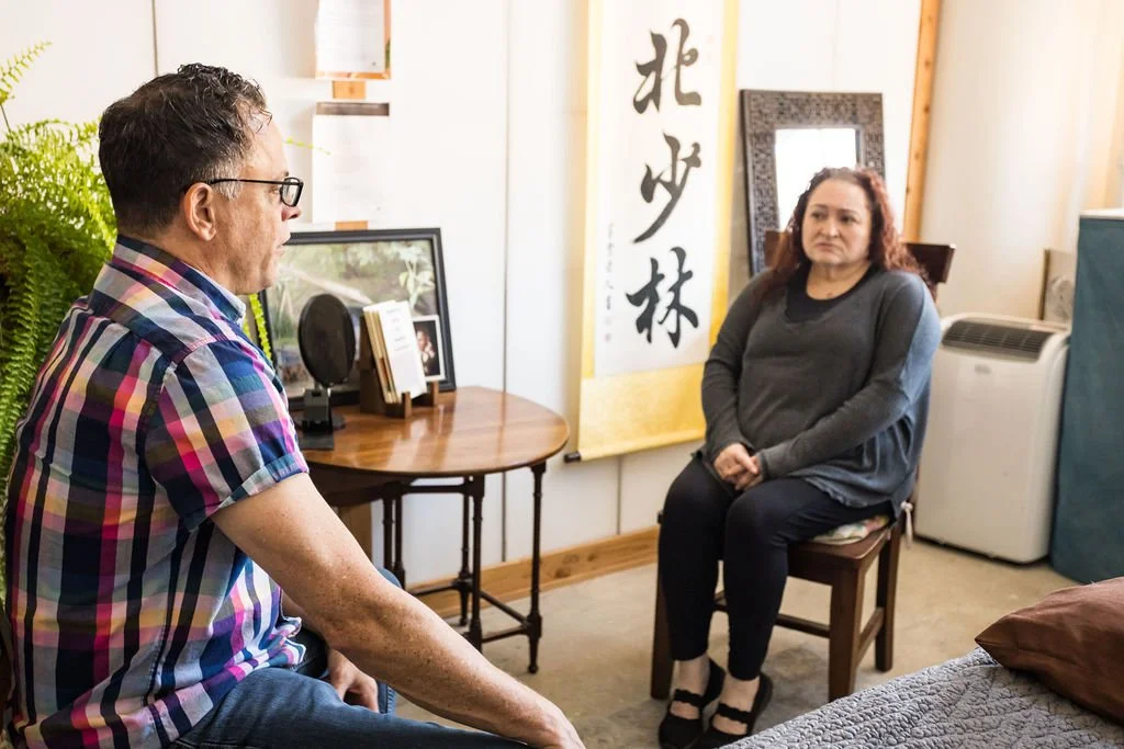 A man and woman sitting in a room engaged in a conversation. The man, on the left, wears glasses and a colorful plaid shirt. The woman, on the right, has curly hair and wears a gray sweater. The room has a table with framed pictures and a plant, and a large Asian calligraphy scroll in the background.
