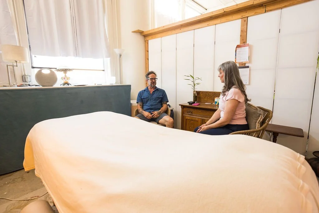 A man and a woman sitting and talking in a bedroom with a bed in the foreground, a window with curtains, and wooden paneling on the wall.