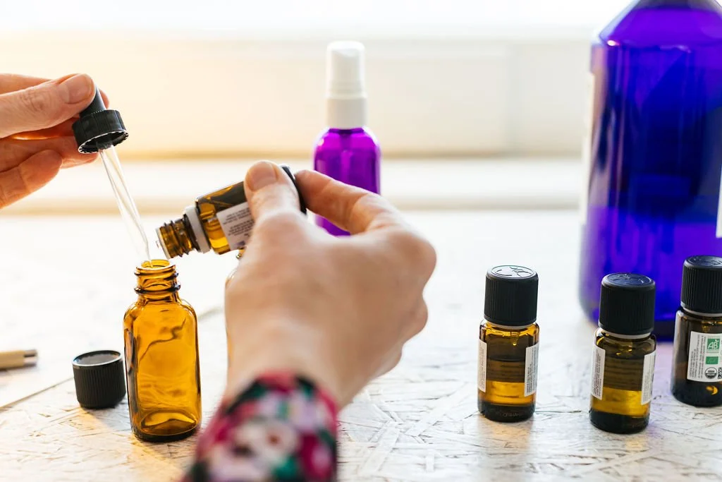 Person adding drops from a small amber glass bottle into an open amber glass bottle, with additional small amber bottles and larger bottles on the table.