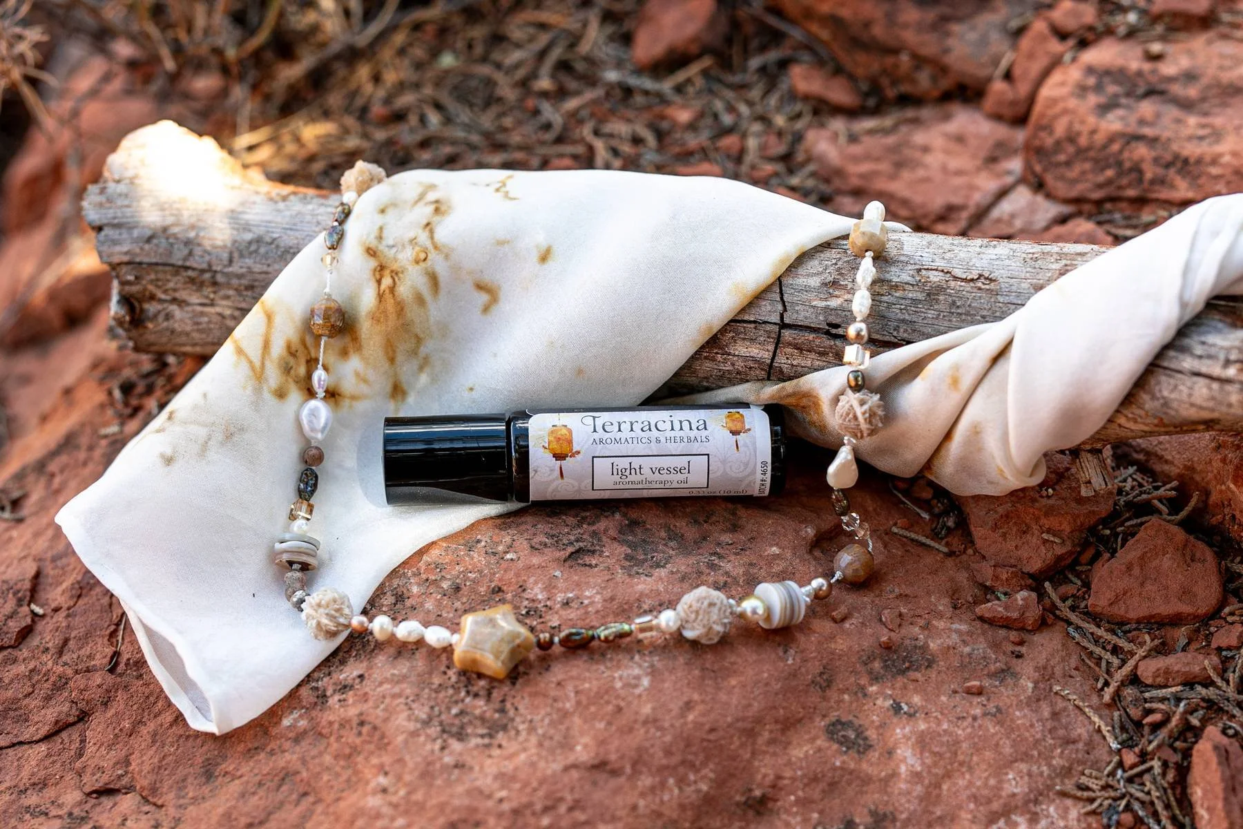 A small bottle of terracina aromatherapy oil labeled 'Light Vessel' lying on a rocky surface, draped with a pearl necklace and a piece of white fabric.