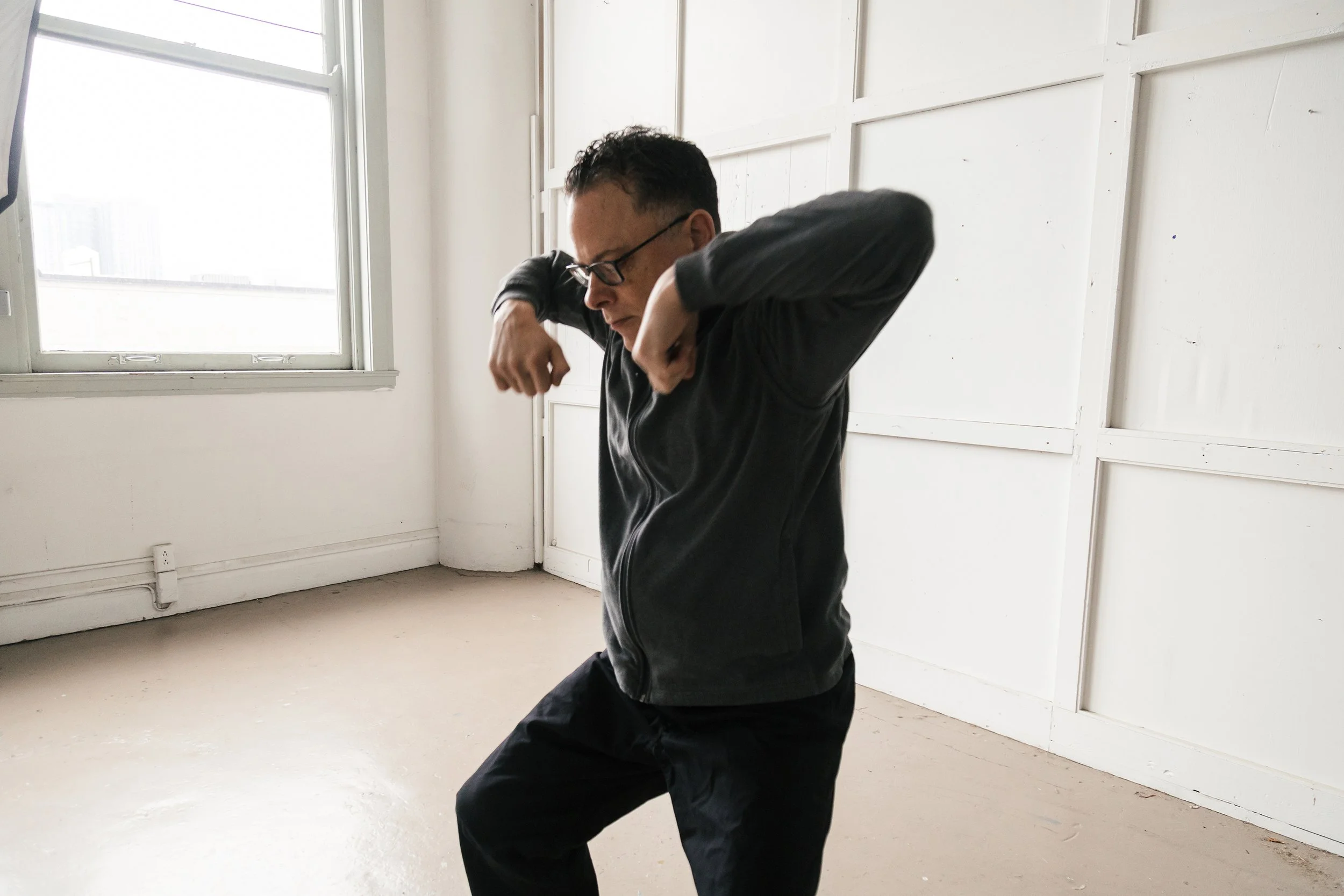 A man wearing glasses and black clothing is practicing a martial arts pose indoors, with white walls and a window in the background.