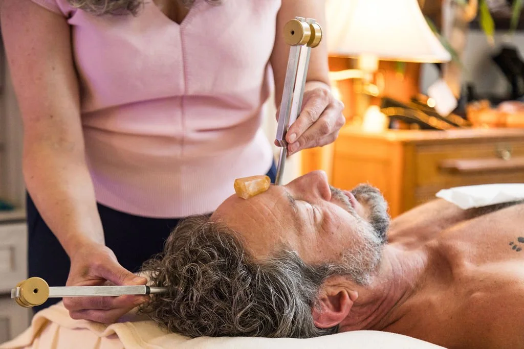 A person lying on a bed receives an at-home crystal healing treatment from another person using metal and crystal tools.