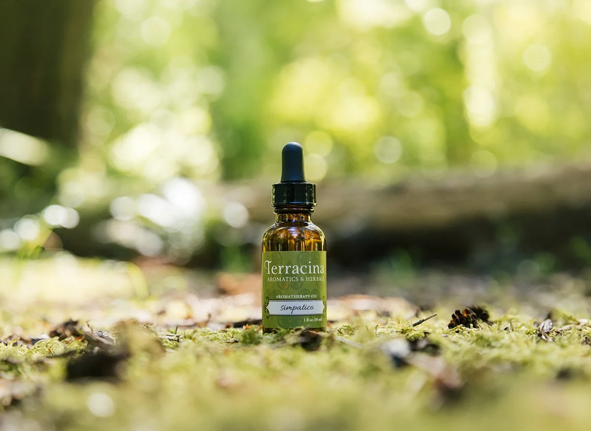 A small amber glass bottle with a black dropper cap placed on a forest floor covered with moss and small leaves, with blurred green trees in the background.