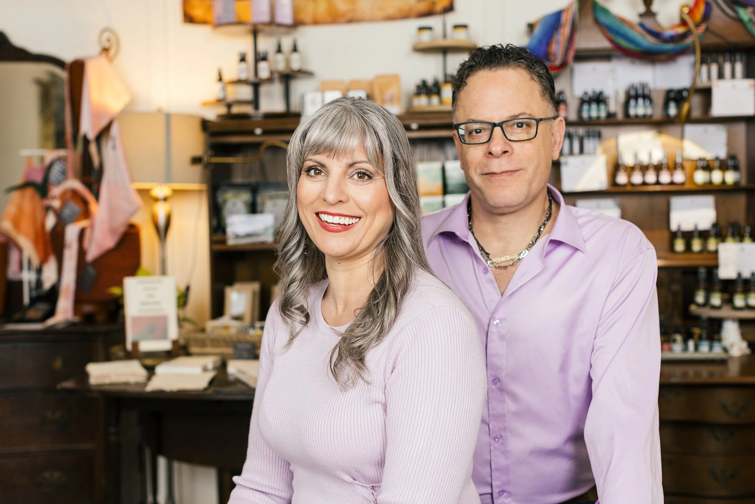 A smiling woman with gray hair and a man with glasses in a store with shelves of bottles and packages in the background.